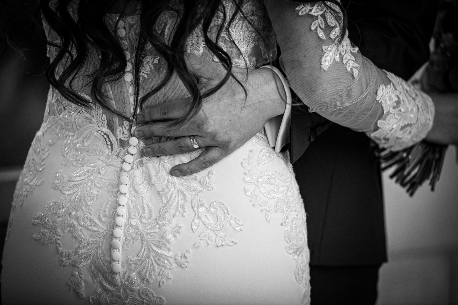 A black and white detail shot showing wedding dress with intricate lace patterns and the groom's hand with ring