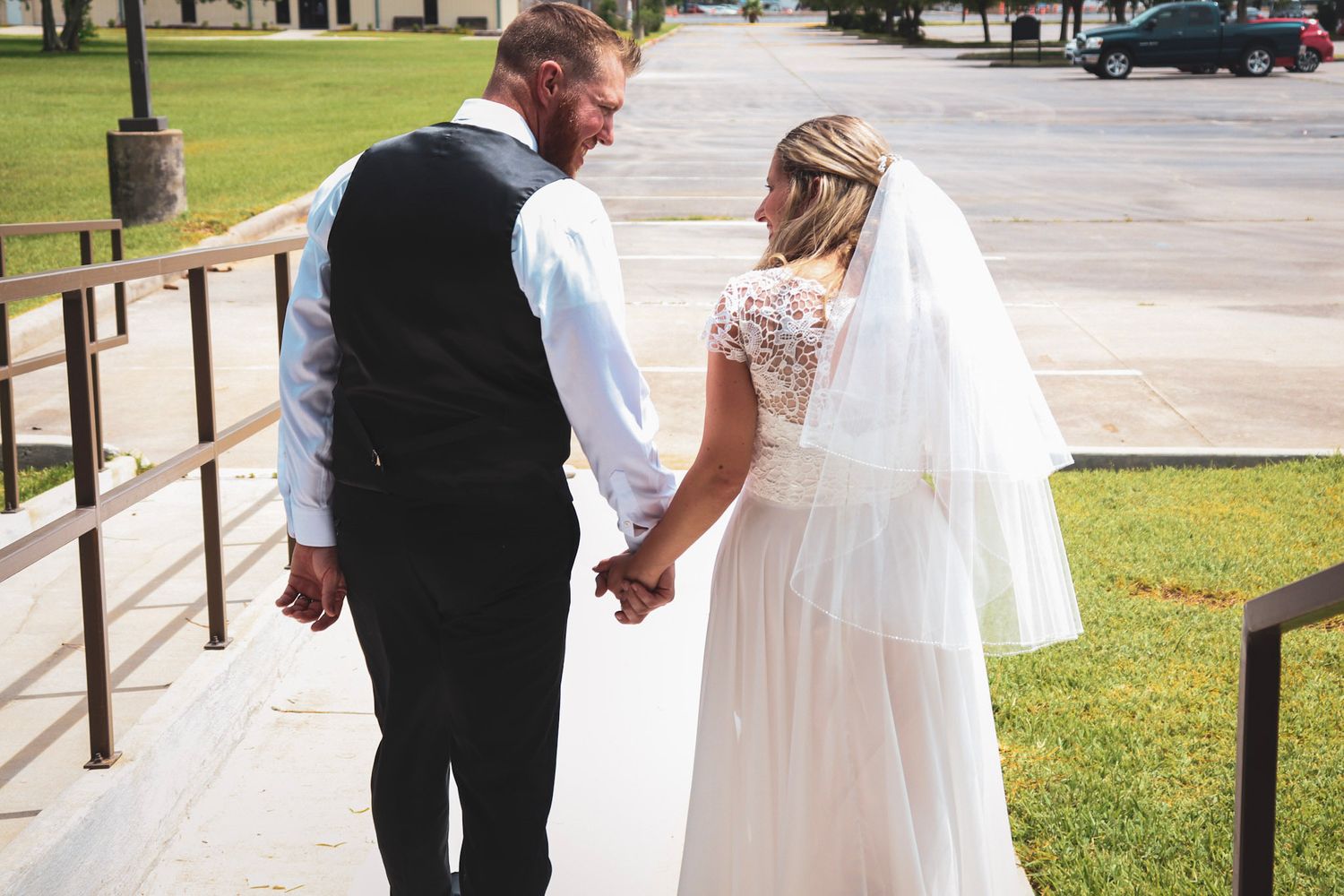 A wedding couple walks along a wooden railing pathway surrounded by green grass and trees on a sunny day.