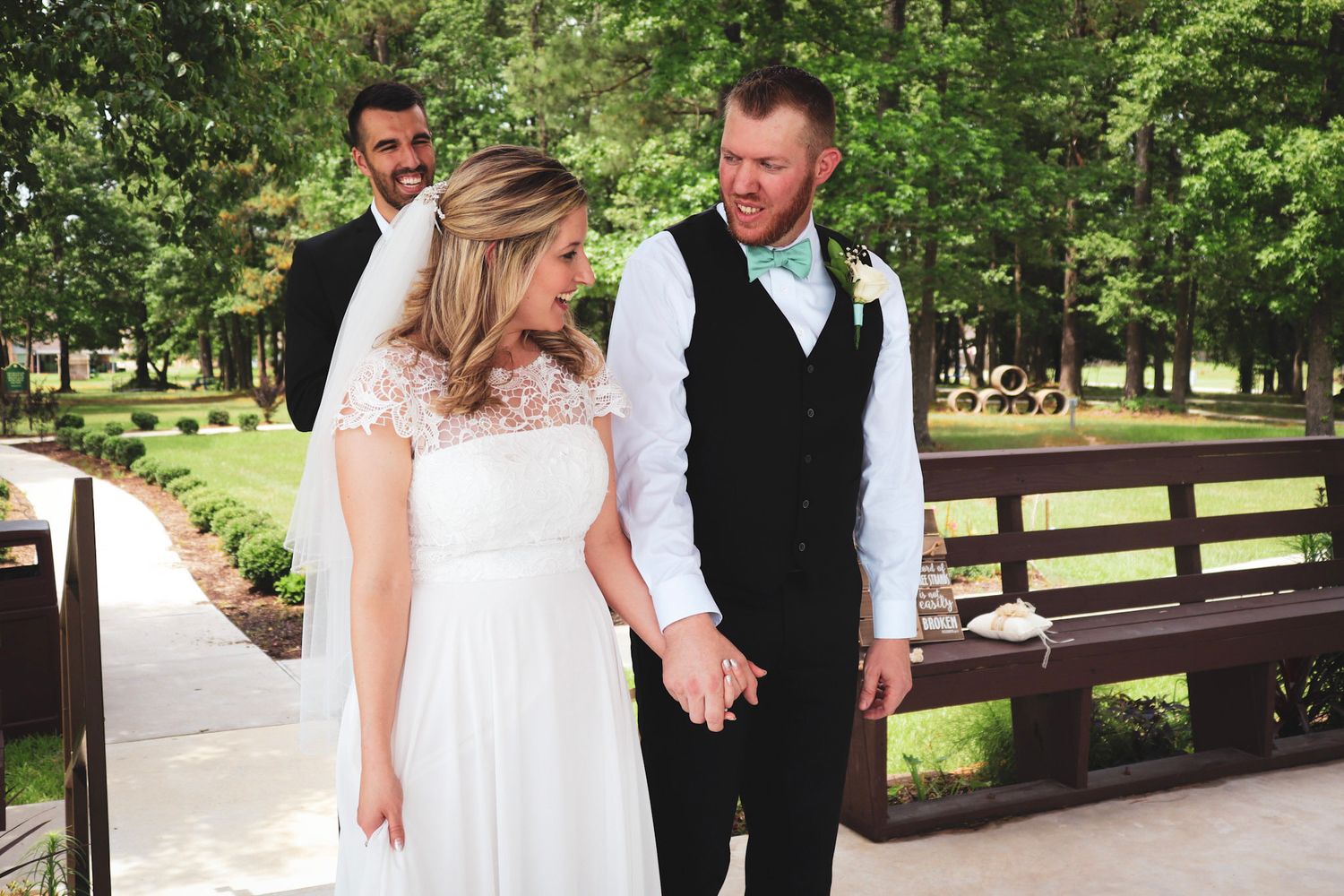 A wedding party stands on a wooden bridge in a lush green park setting during a summer celebration.