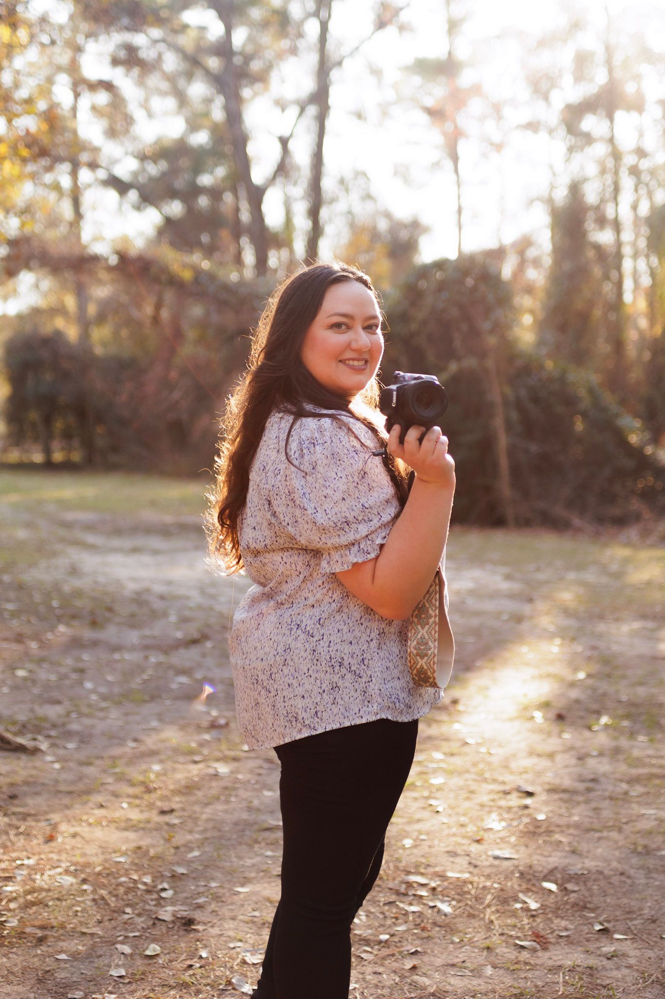 Photographer with long dark hair captures sunset photos in an autumn forest setting wearing a floral top and black pants.