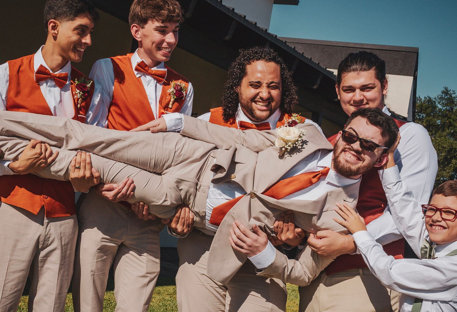 Groomsmen in khaki pants and orange vests playfully pose together outdoors on a sunny day.