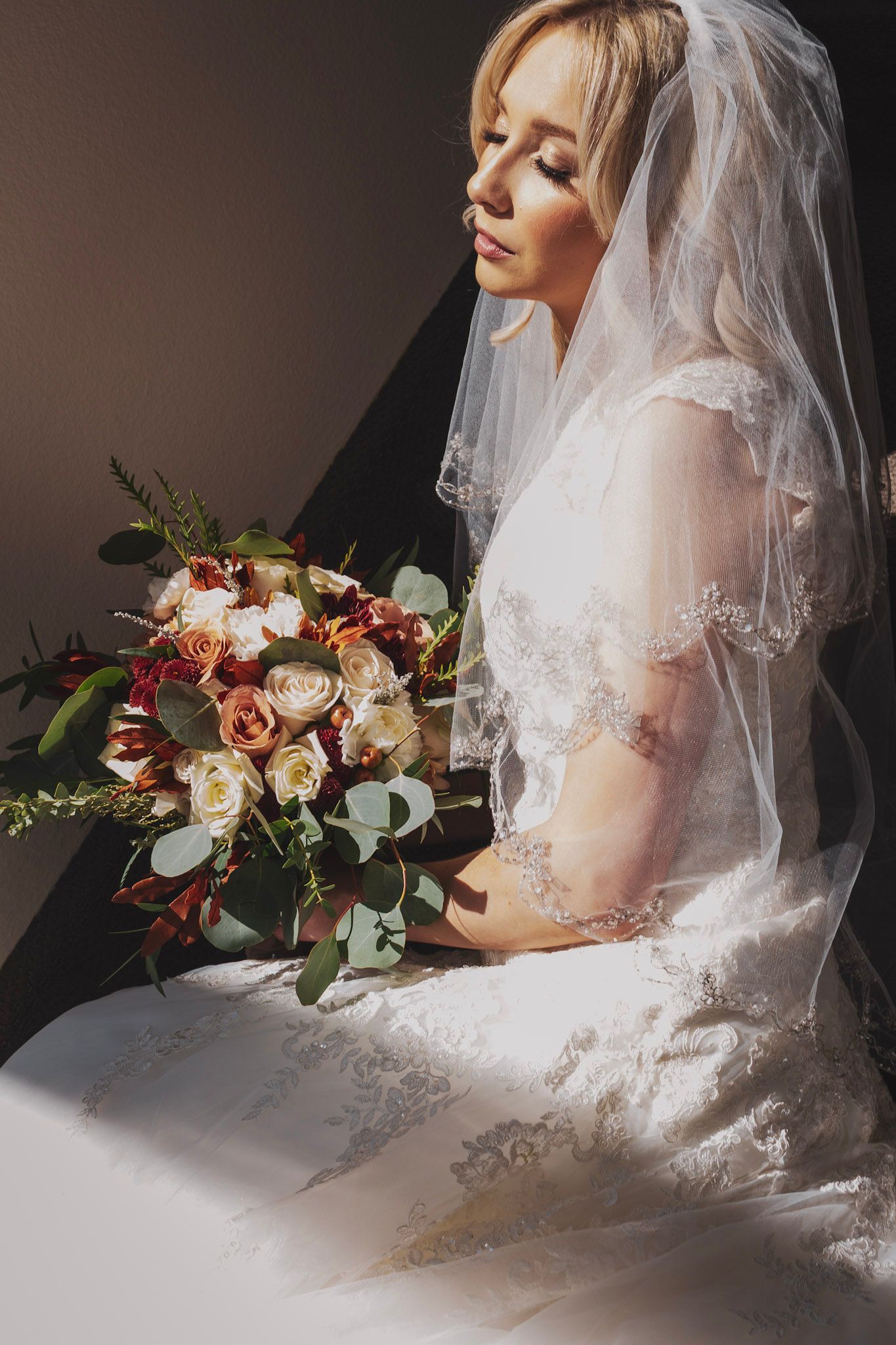 A bride in a lace wedding dress holds an autumn-toned bouquet with roses and eucalyptus against a dark background.