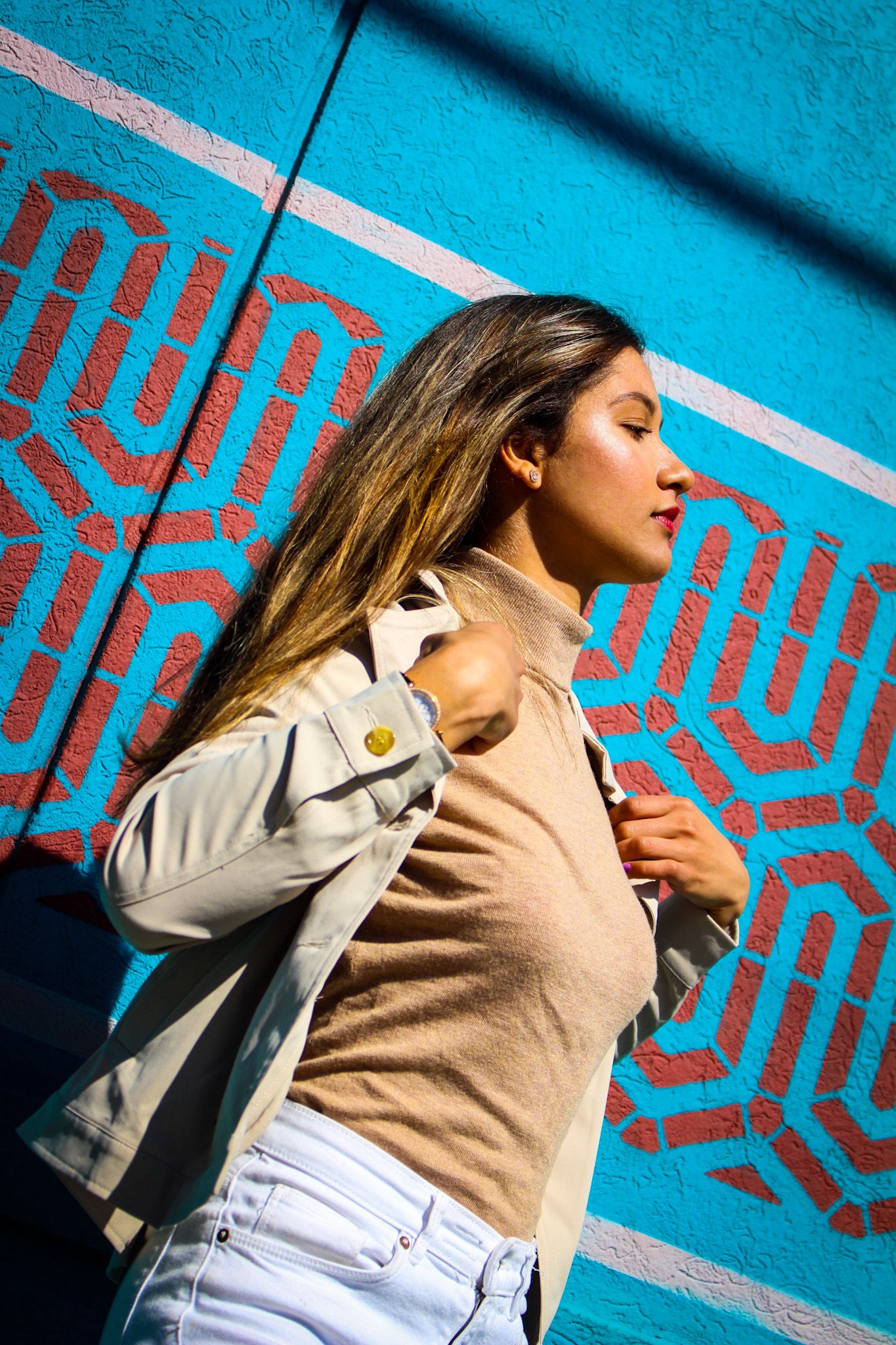 Person in beige sweater and light jacket poses dramatically against vibrant turquoise wall with red geometric pattern.