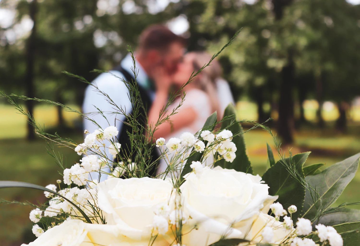 Beautiful white wedding bouquet with roses and greenery in focus while couple embraces in blurred background outdoors.