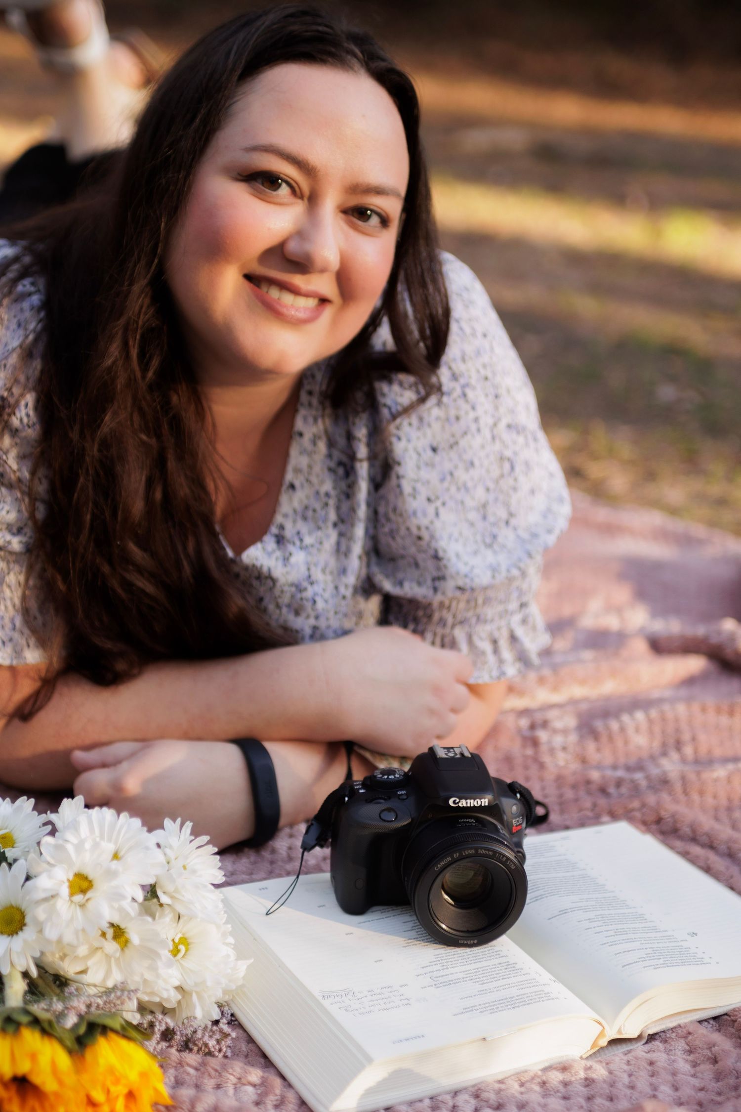 A person lies on a blanket outdoors with a Canon camera, open book, and fresh daisies on a sunny afternoon.