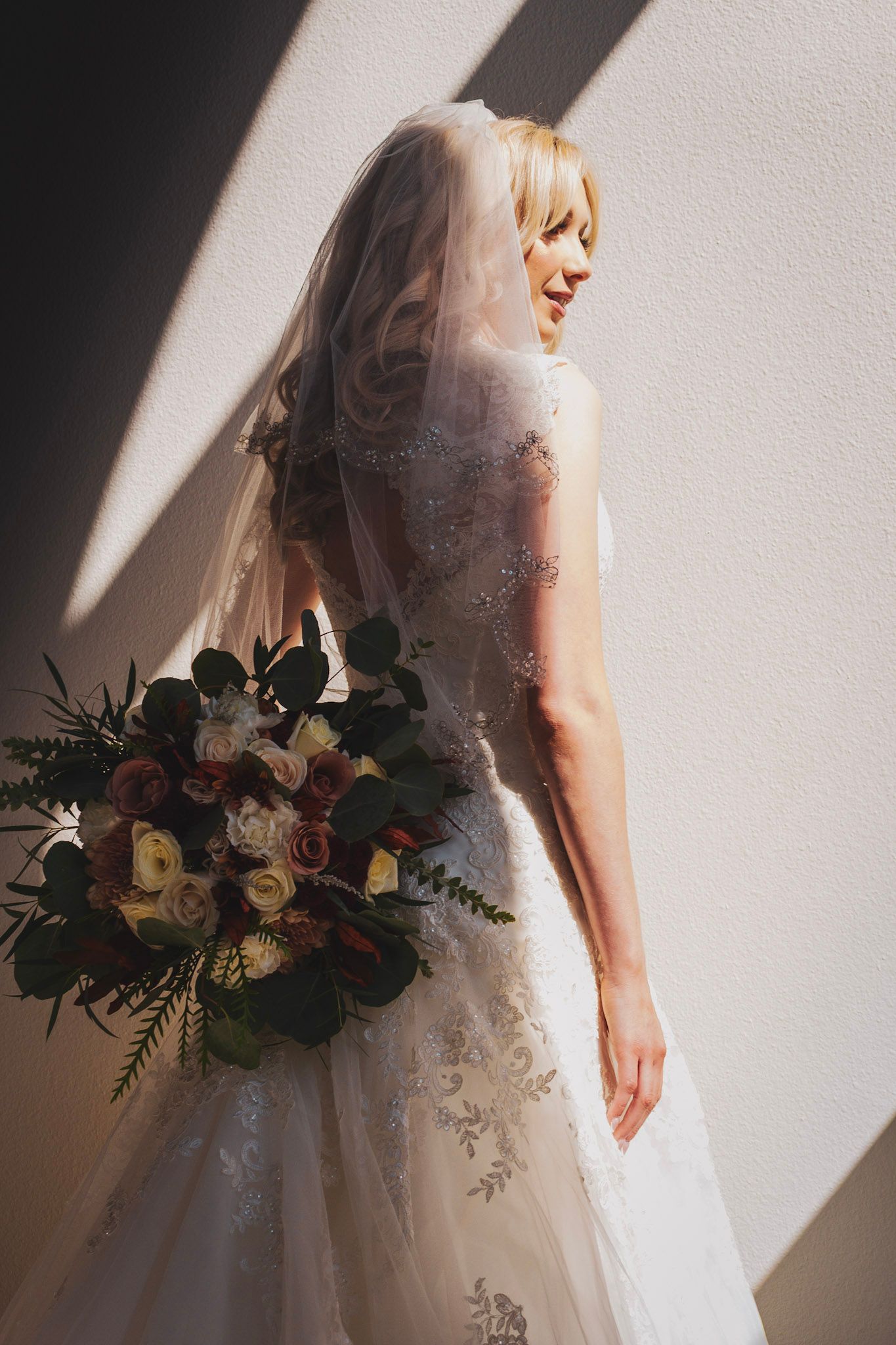Bride in white lace wedding gown holds elegant burgundy and blush floral bouquet in dramatic window lighting.