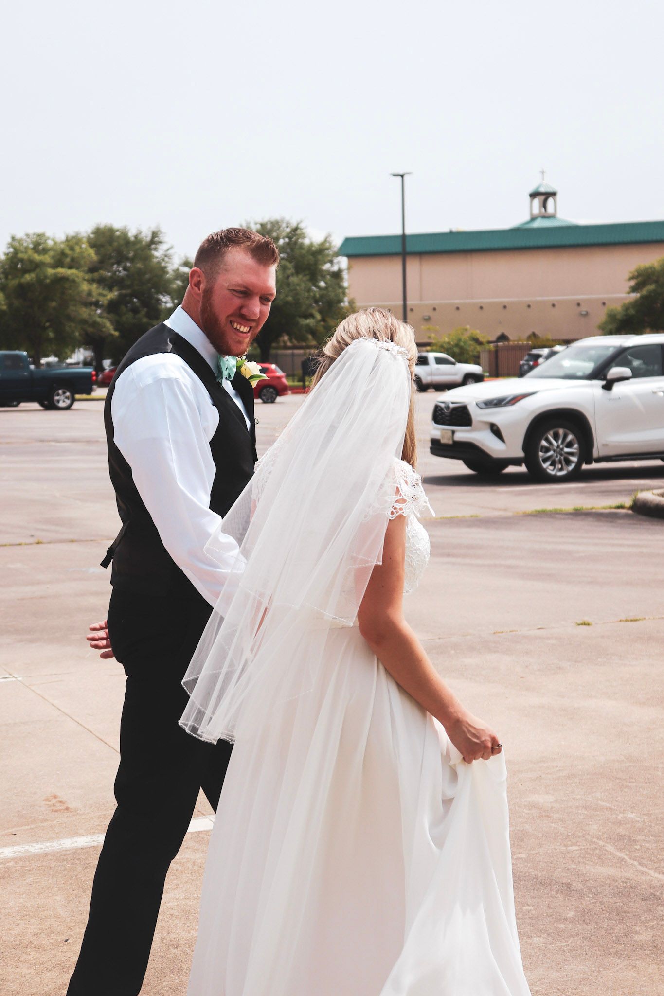 A couple in wedding attire share a joyful moment in a parking lot, with the bride wearing a flowing white veil.