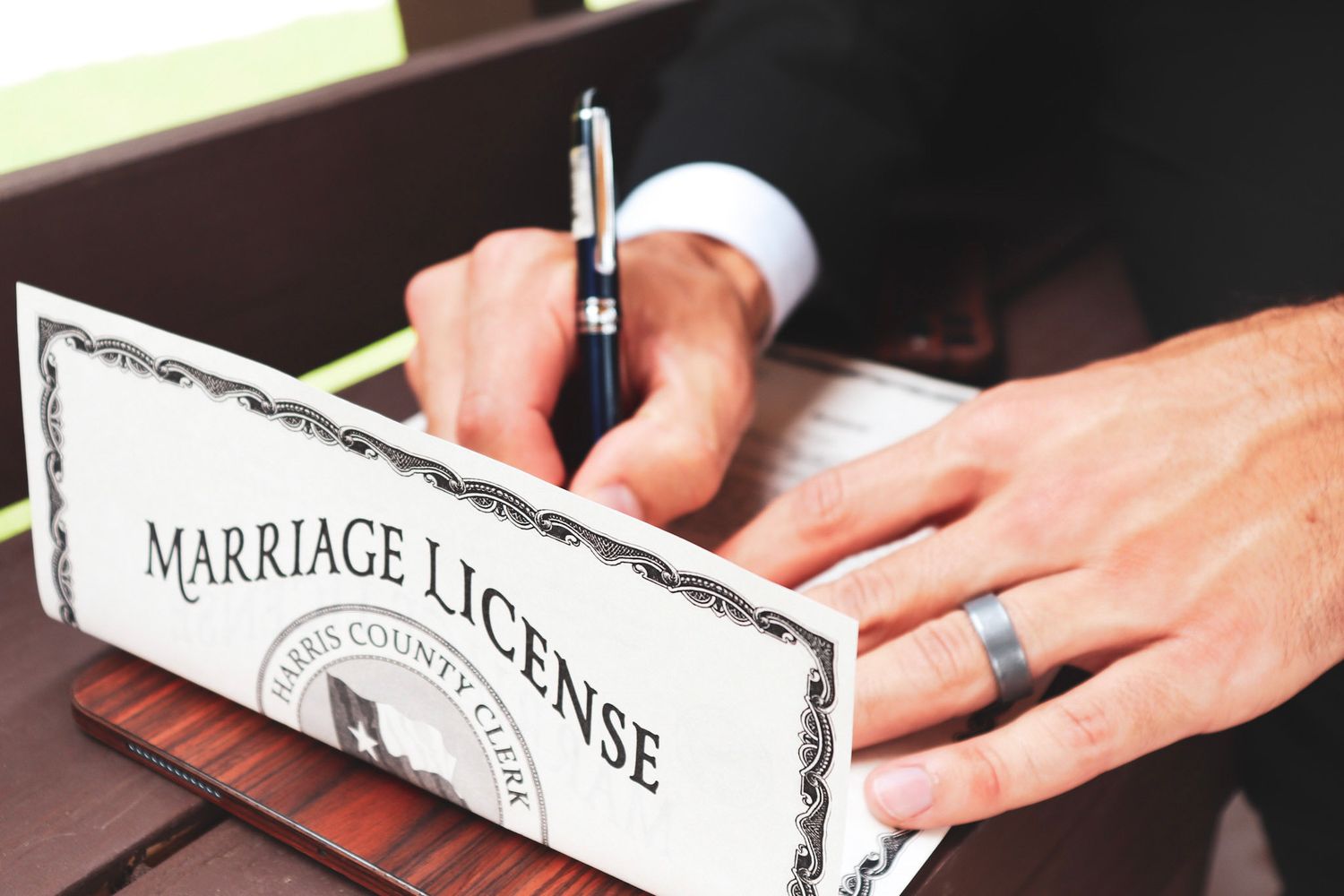 Hands signing a marriage license document with a pen on a wooden table surface.