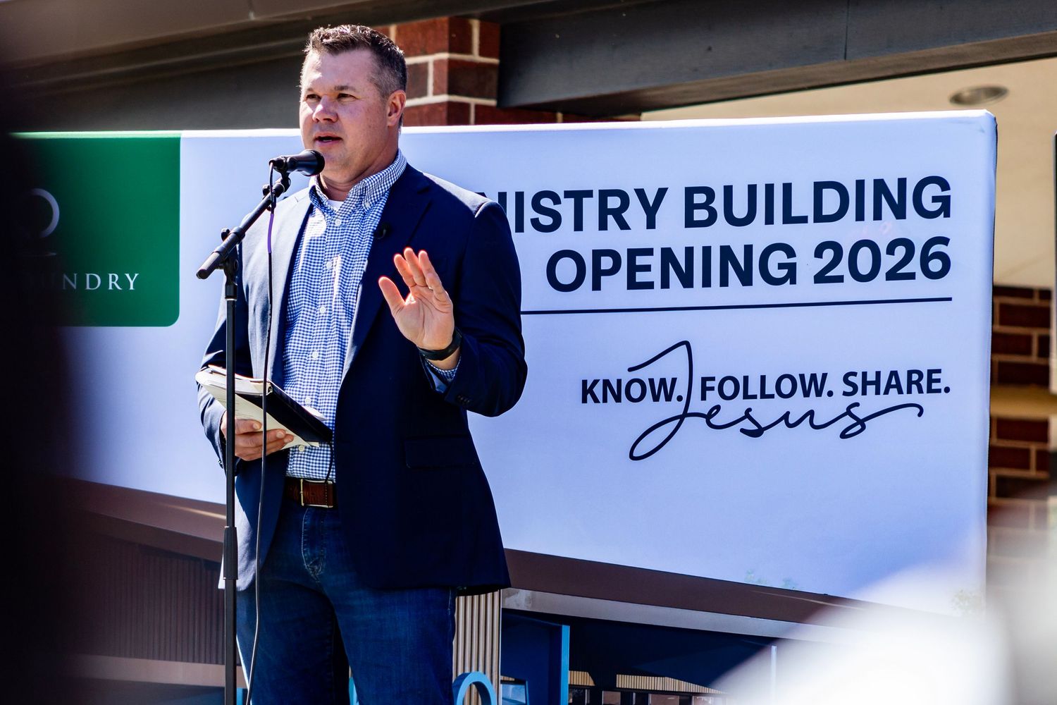 A speaker in a navy blazer gestures while presenting in front of a banner announcing a Ministry Building opening in 2026.