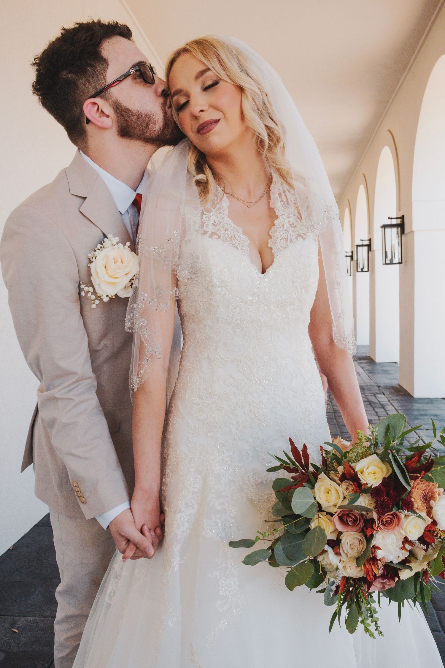 A romantic wedding couple embraces in a light-filled hallway with a beautiful burgundy and cream floral bouquet.