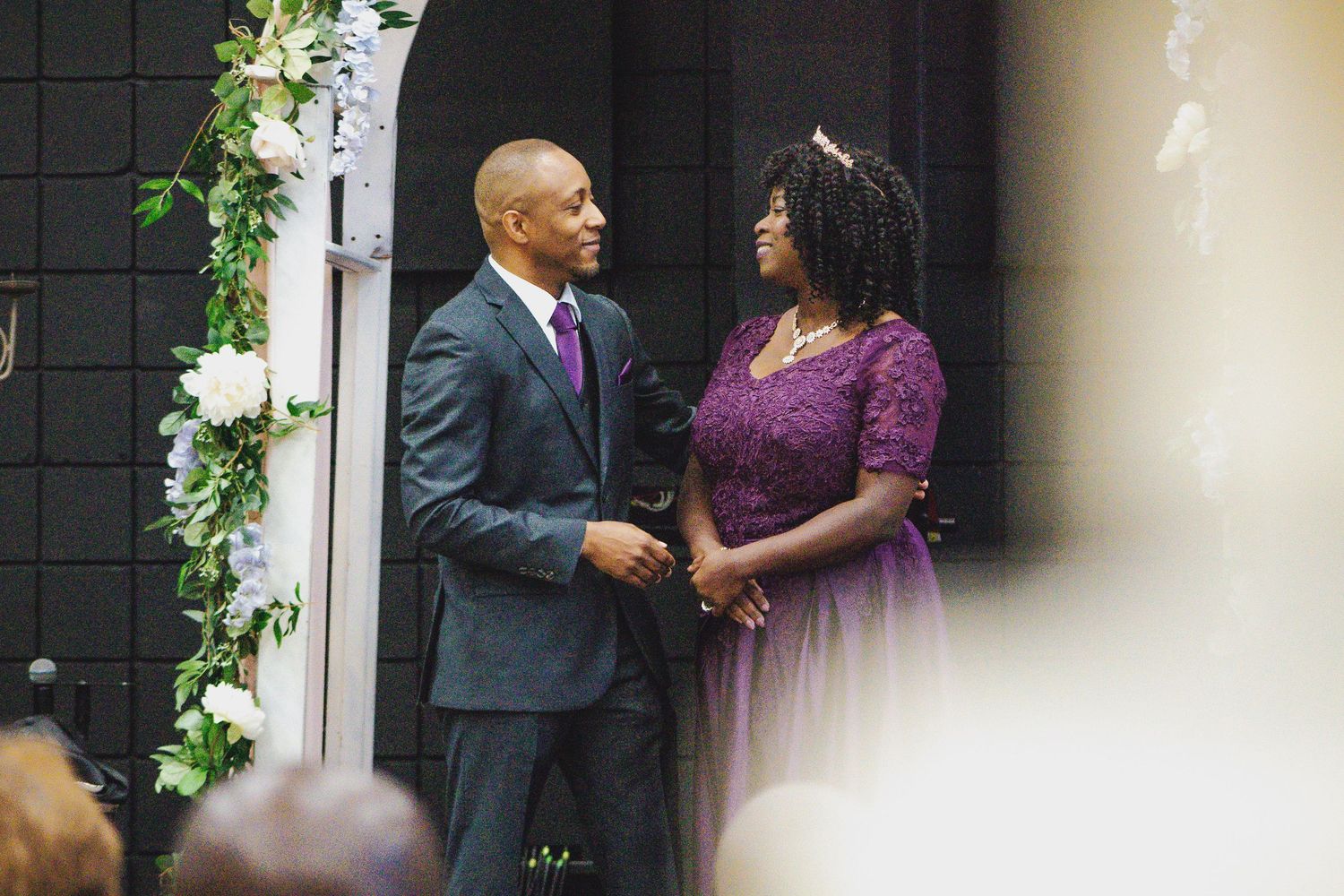 A couple shares an intimate moment in formal attire against a garden backdrop with white flowers and greenery.