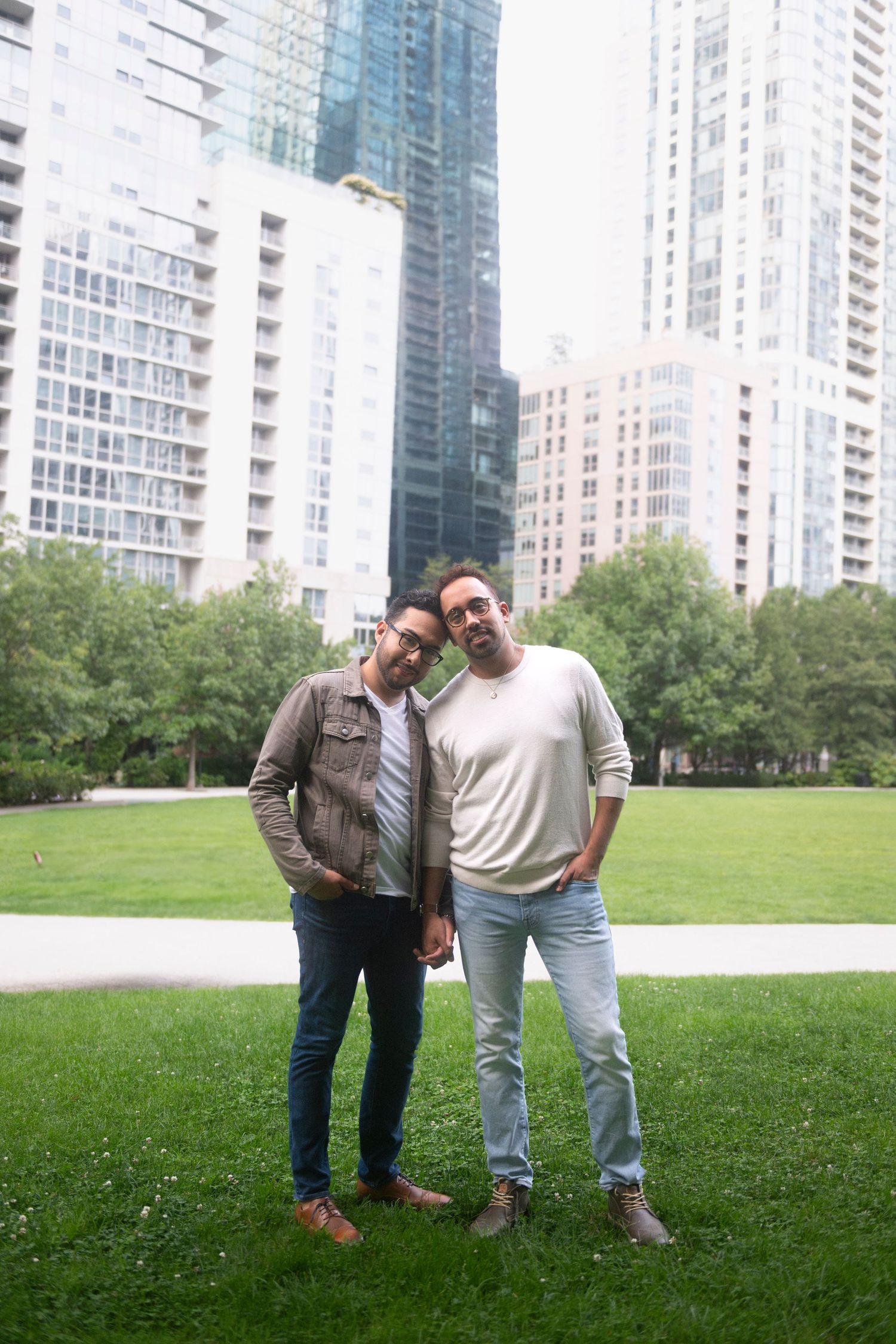 Two men hold hands and stand together, resting their temples against each other, and look at the camera.