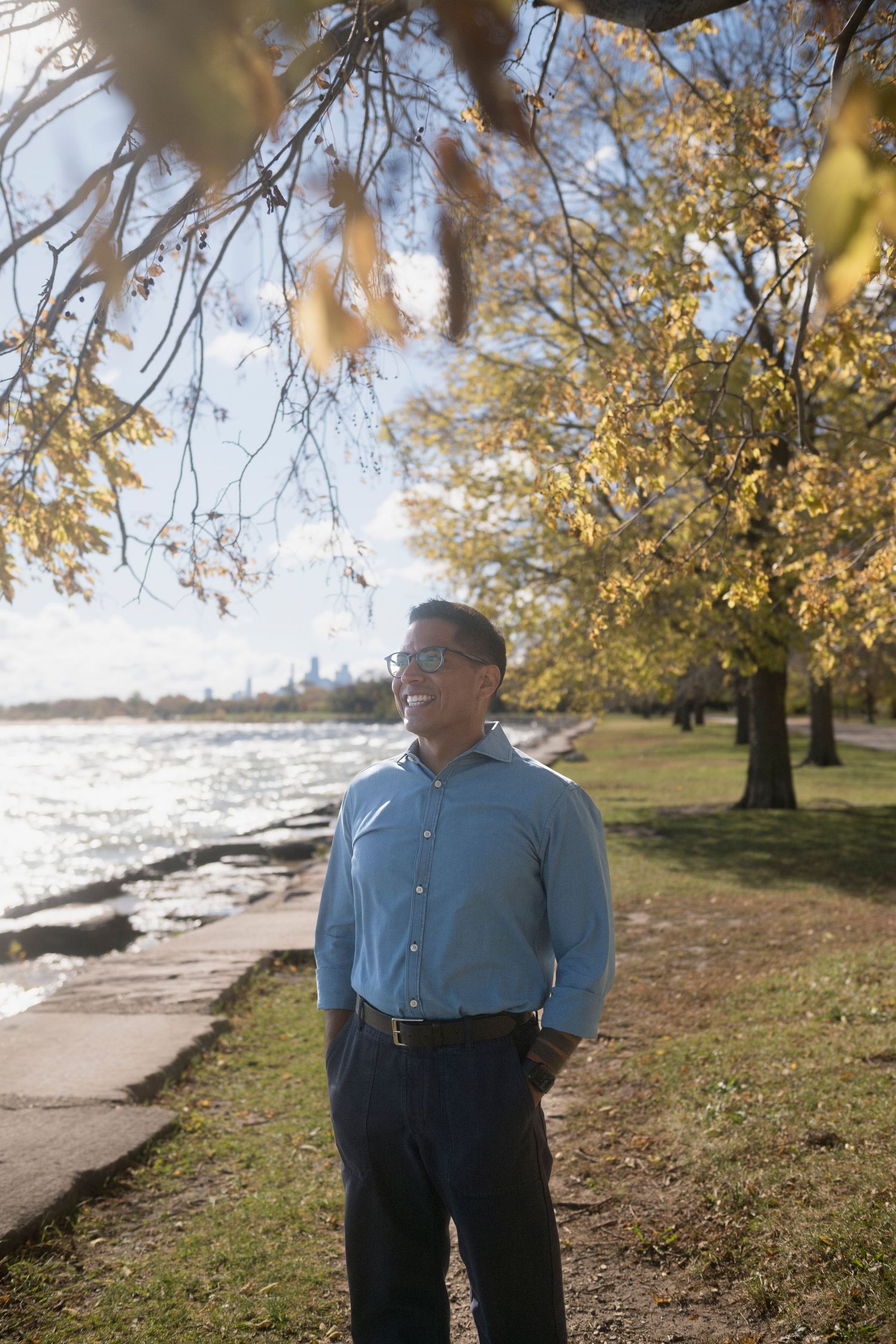A man stands among the trees along the lakefront with his hands in his pockets, smiling out over the water on a bright and sunny day.
