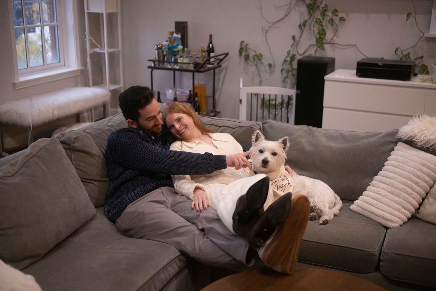 A newly engaged couple snuggle on the couch in their living room with their white West Highland Terrier.