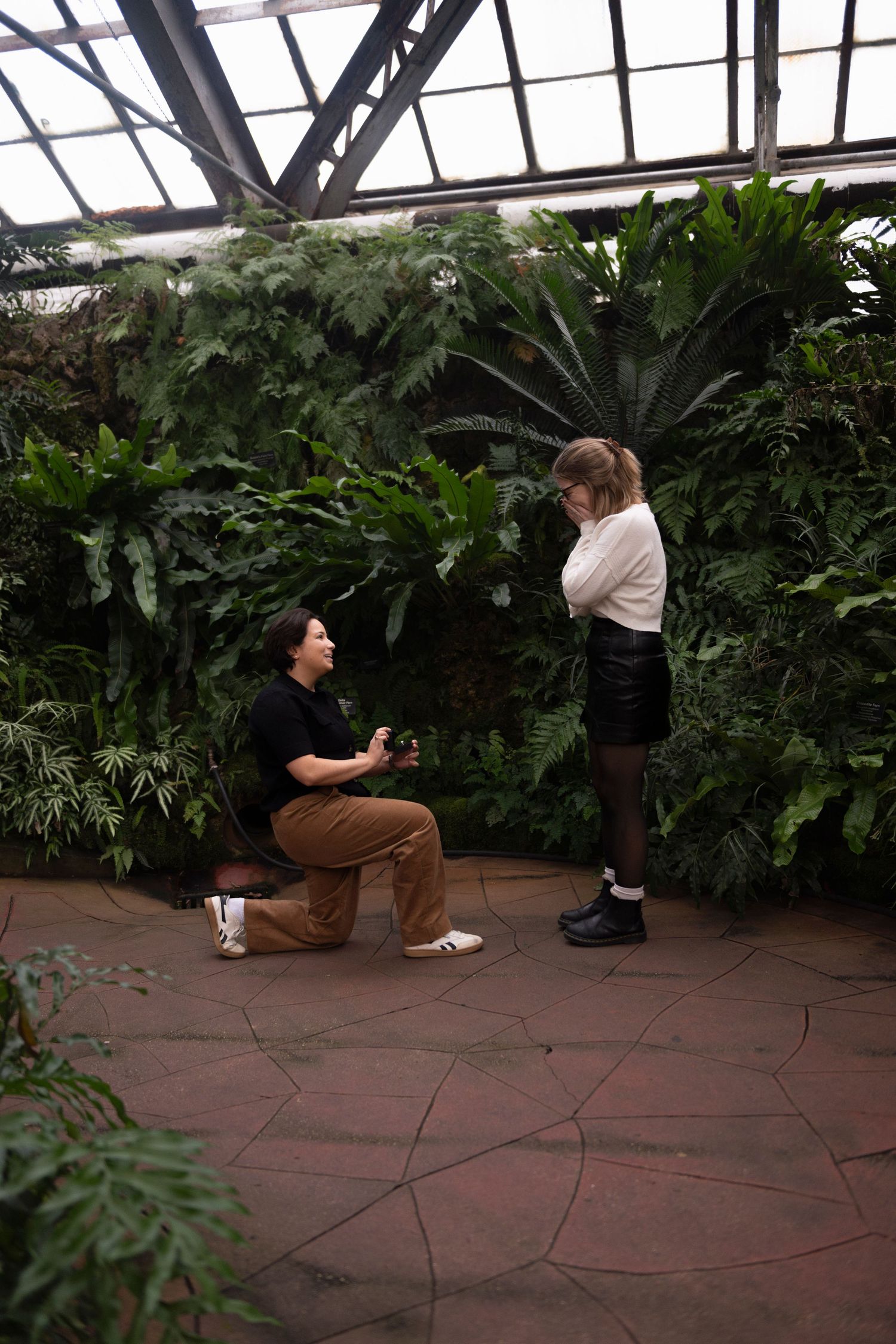 A young woman kneels with a ring box in front of her girlfriend, who has her hands covering her mouth in surprise.