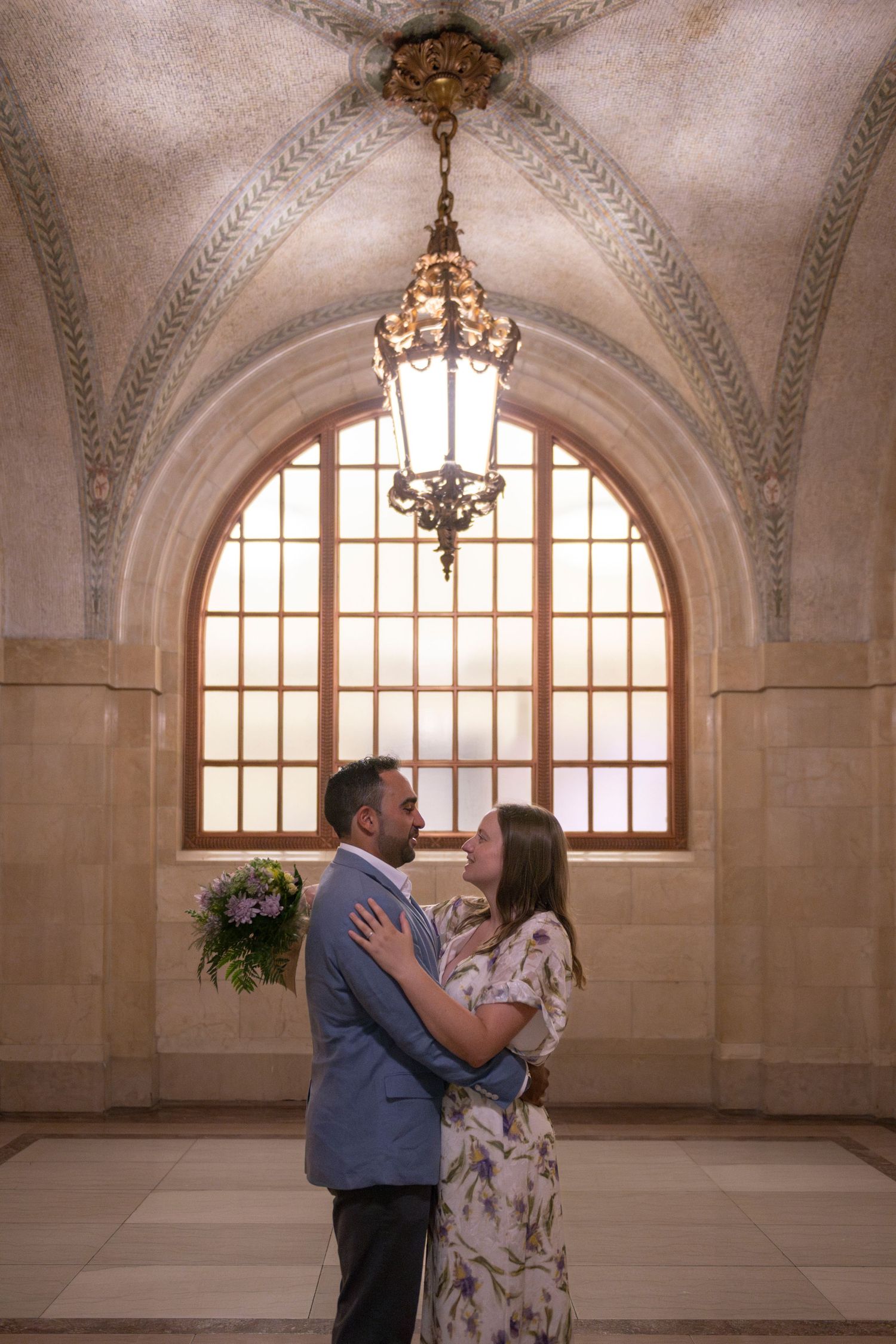A couple face and embrace each other under archways and a chandelier inside Chicago's City Hall (vertical orientation).