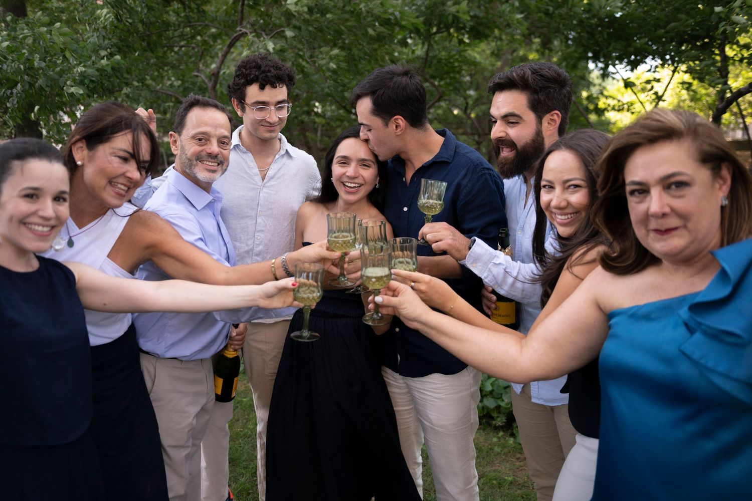 A man kisses his fianceé's temple while she smiles and they cheers their family surrounding them, holding champagne glasses.