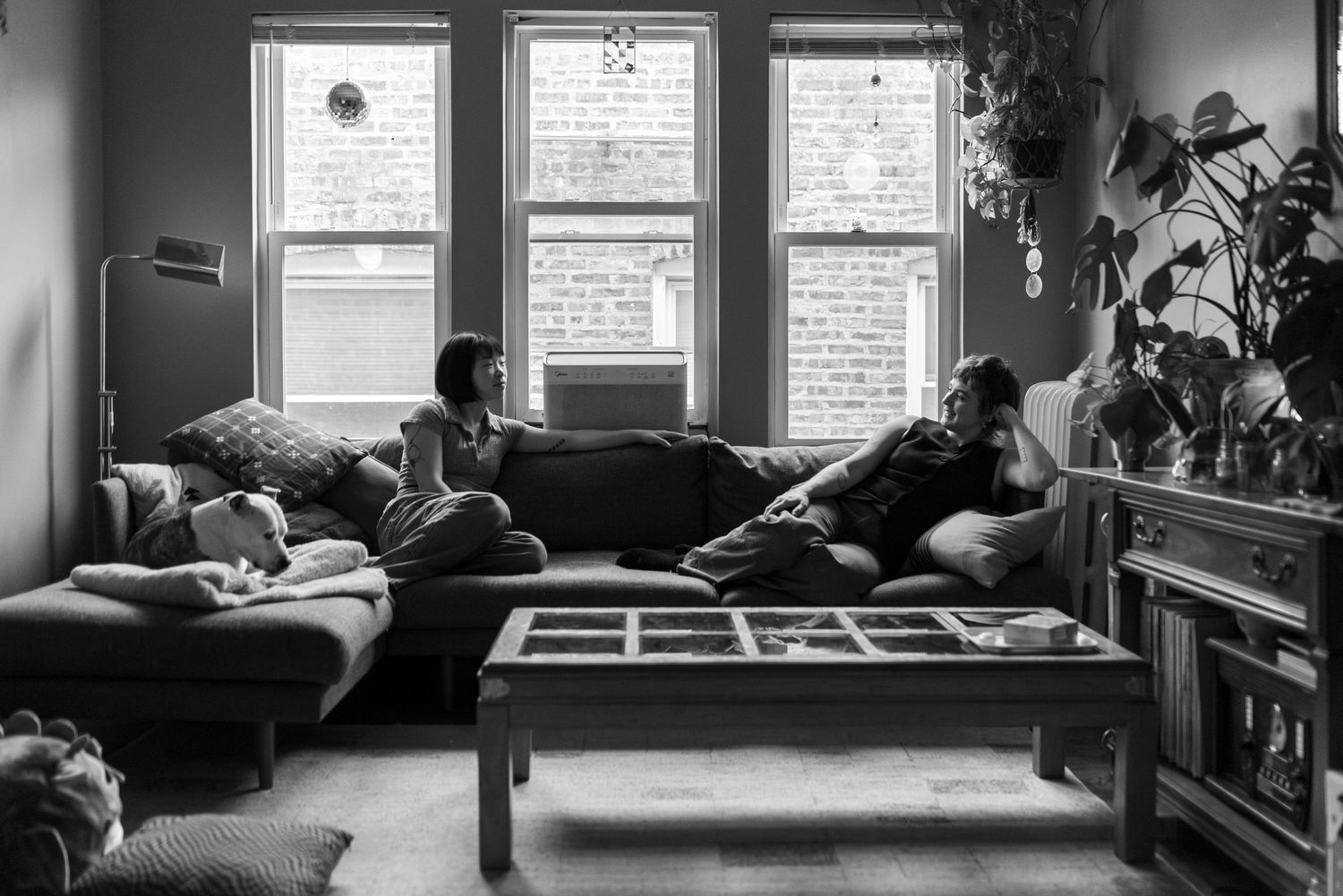 A queer couple sits on their couch in front of windows, relaxed and facing each other, while their dog lays on the end (B&W).