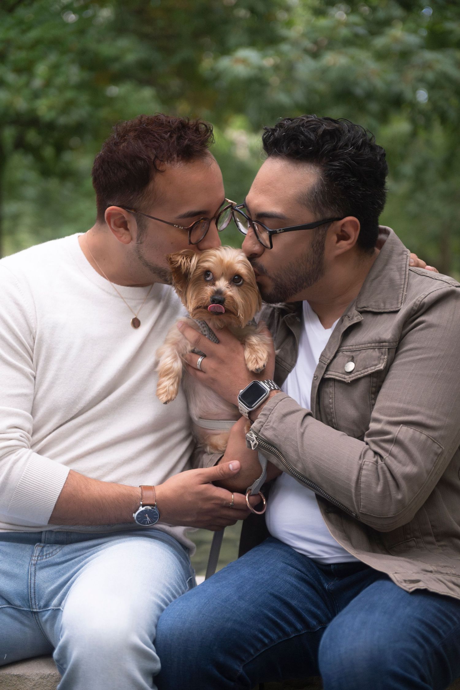 A gay couple hold their Yorkshire Terrier in between them as they sit, and kiss her on the head at the same time.