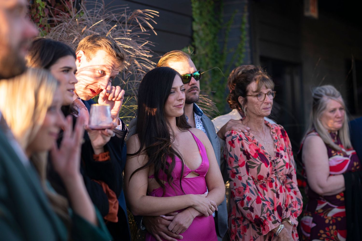 Family and friends stand in the shade and watch their loved ones get married at a rooftop wedding on a sunny day, while wiping away tears.
