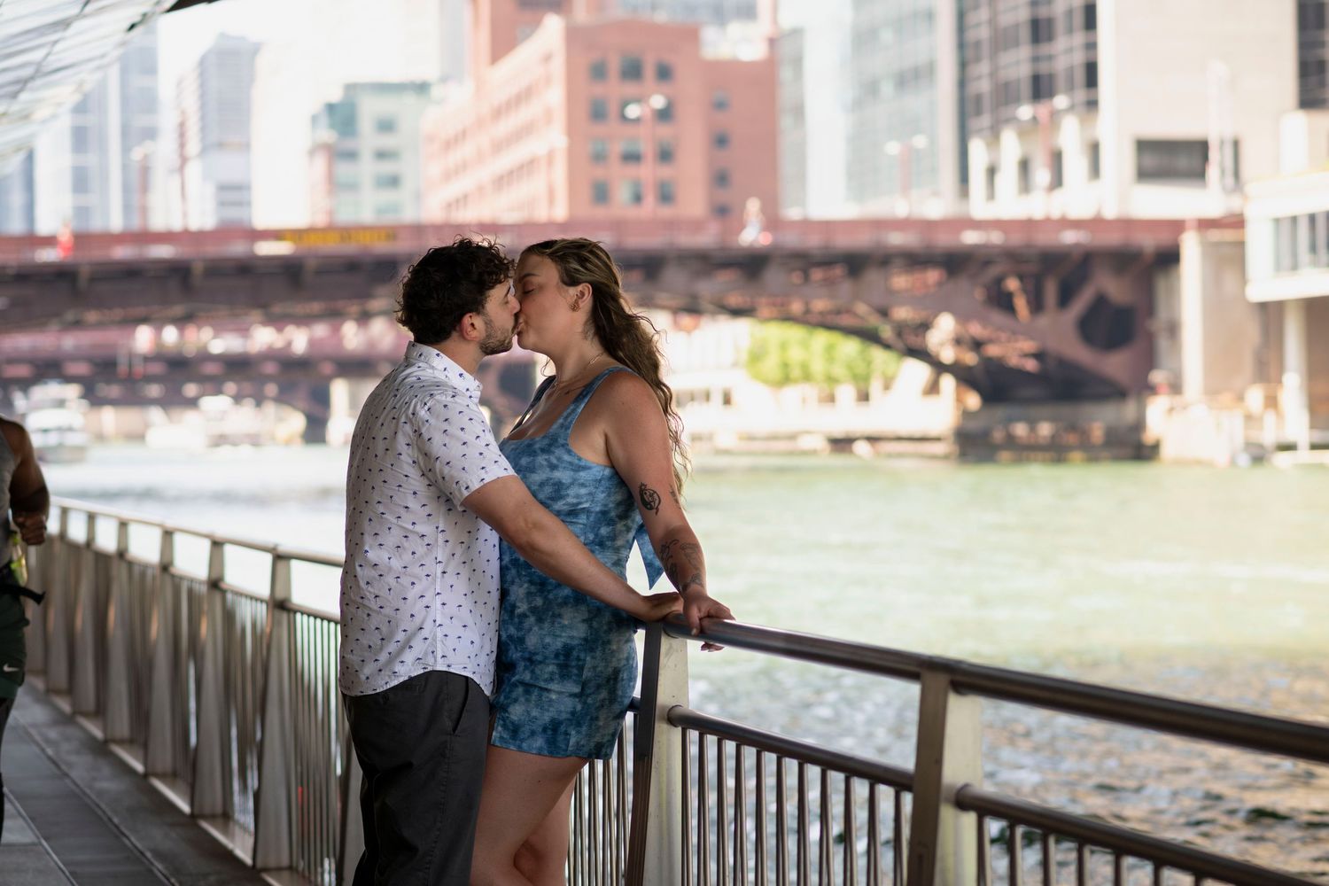A couple kiss underneath a bridge along Chicago's riverwalk on a bright summer day, with a glittering river and bridge in the background.