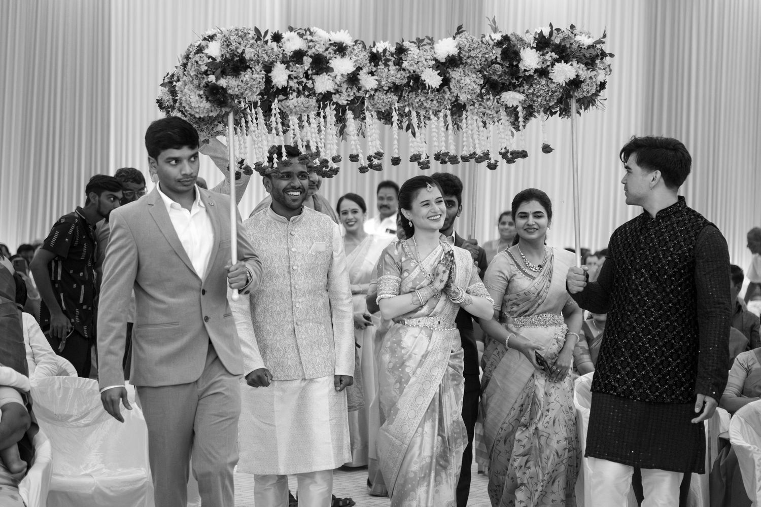 An interracial couple enters their wedding ceremony, underneath a pholoon ki chadar held by both of their male relatives (B&W).