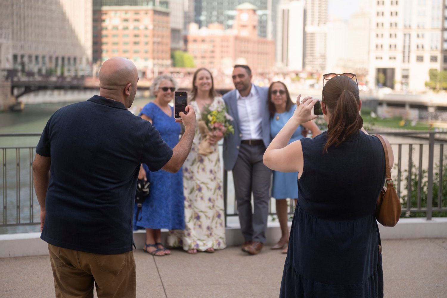 Two people snap photos on their phone of a newlywed couple and their mothers, in front of the Chicago River on a sunny day.