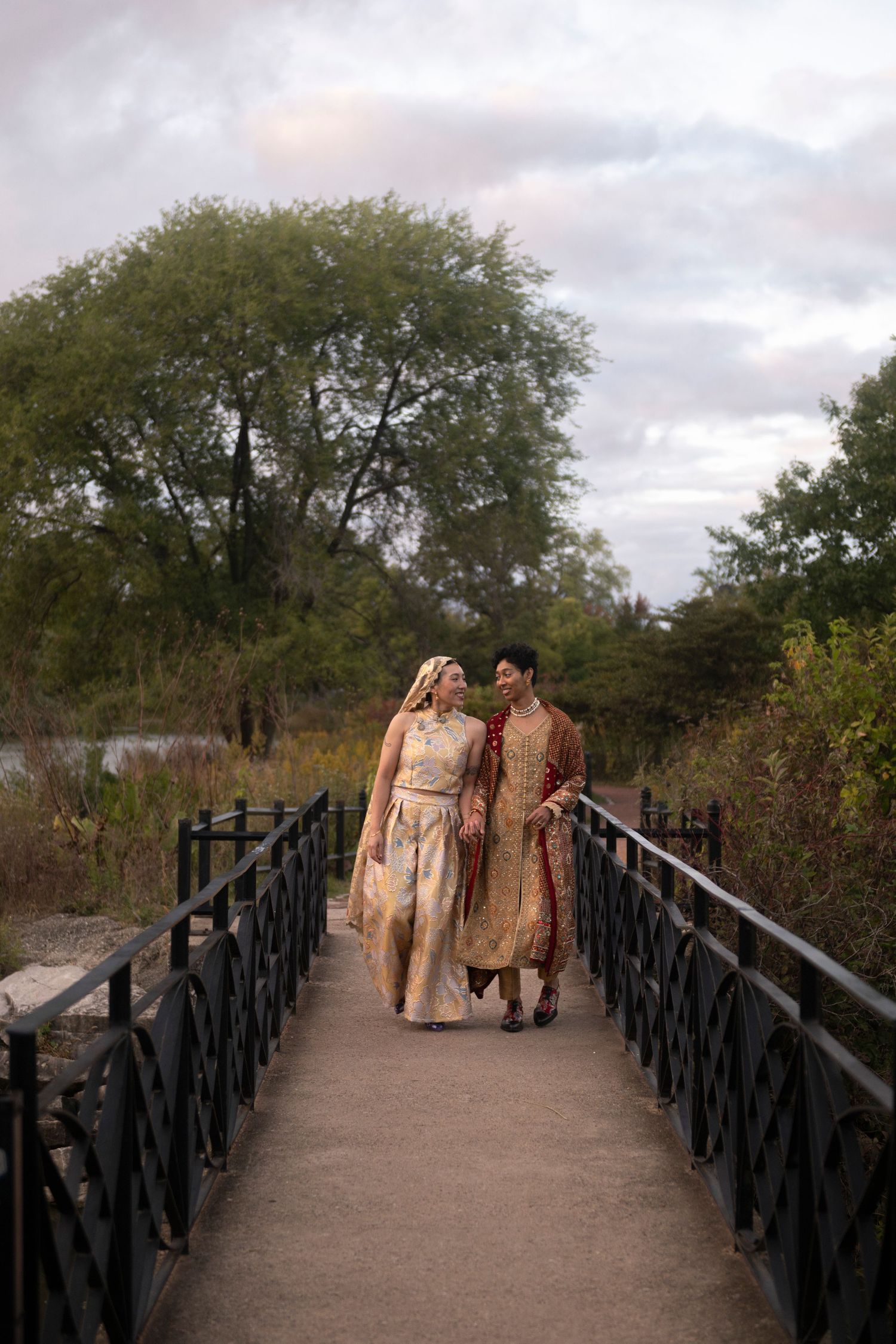 A queer East-and-South Asian newlywed couple walk across a flat bridge over a lagoon at Chicago's Humboldt Park.