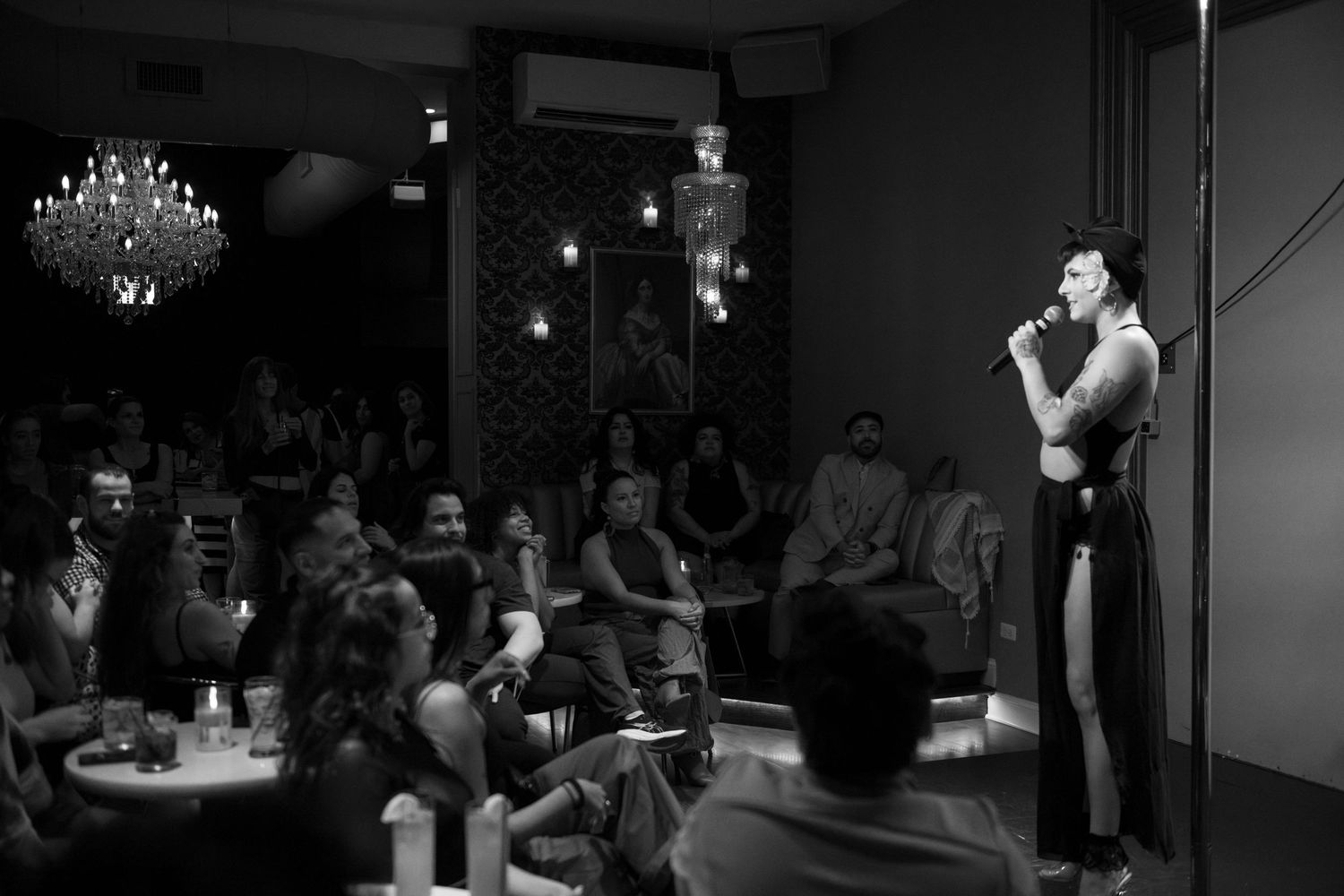 A spotlit performer addresses the crowd from a stage in an intimate pole dancing venue.