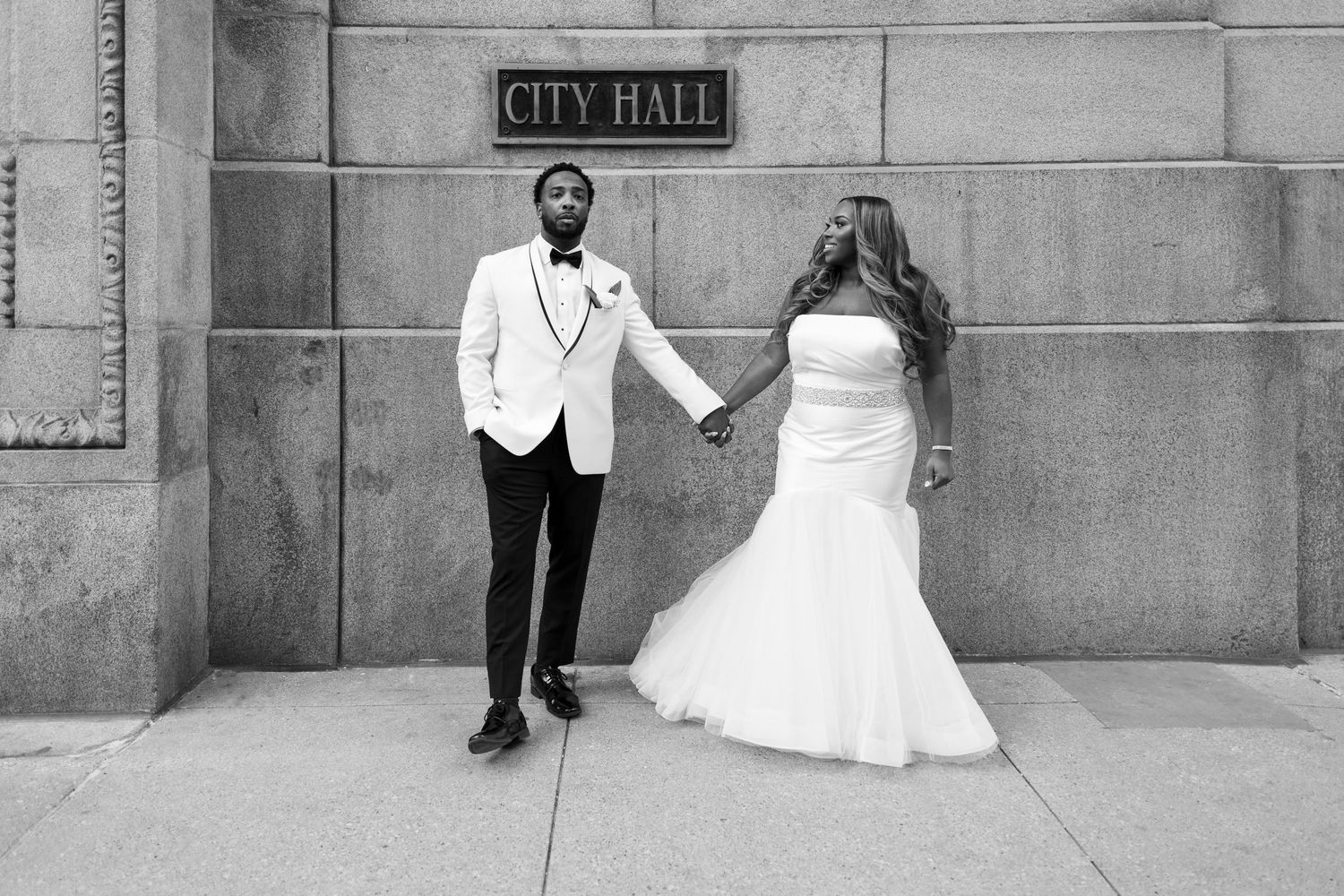 Newlyweds hold hands in front of the exterior City Hall sign in Chicago, Illinois. The bride smiles at her husband while he looks at camera.