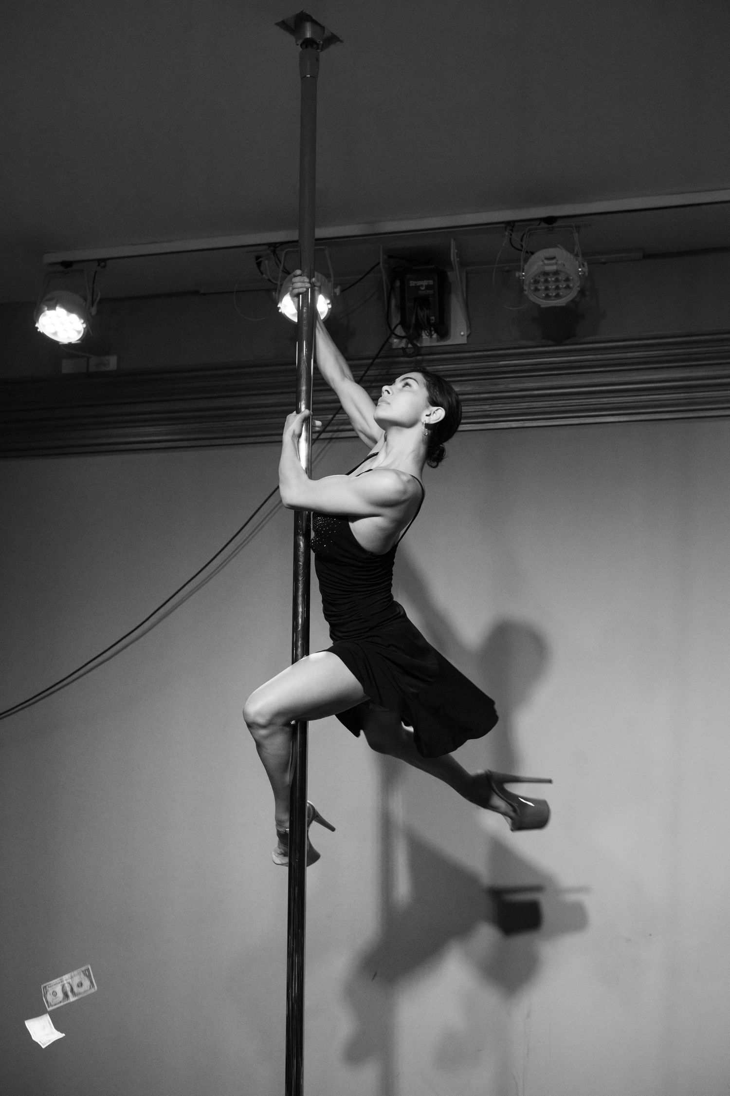 A dancer spins around the pole with one leg hooked around it, while wearing a black mini dress (B&W).