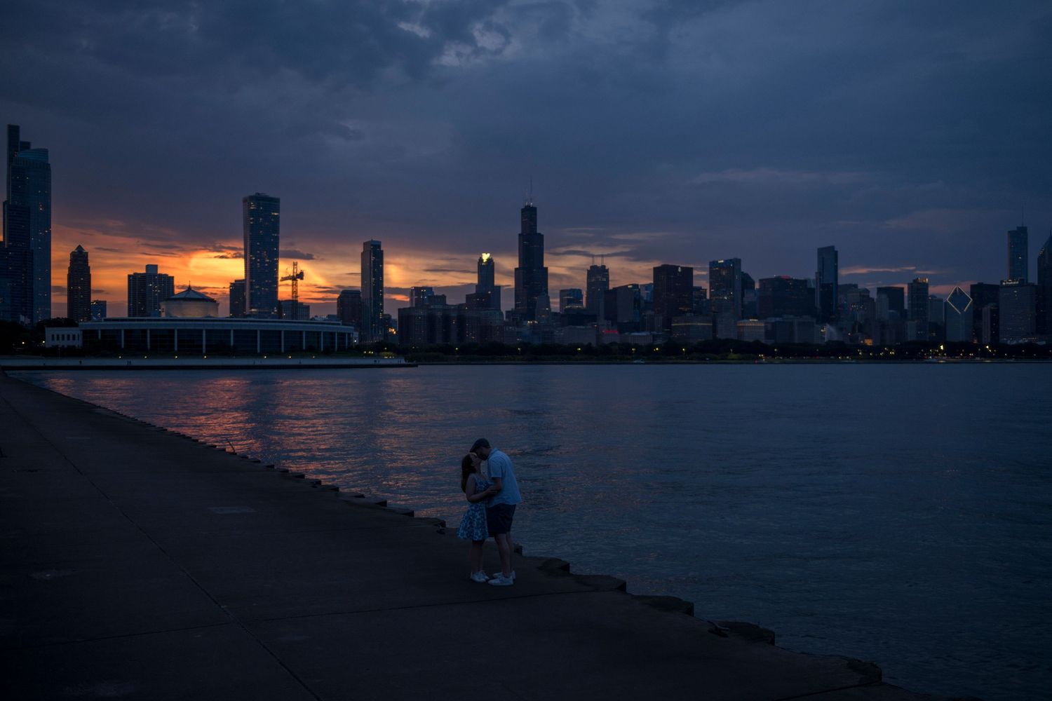 A couple kiss at the beginning of blue hour with the Chicago skyline and Lake Michigan in the background.