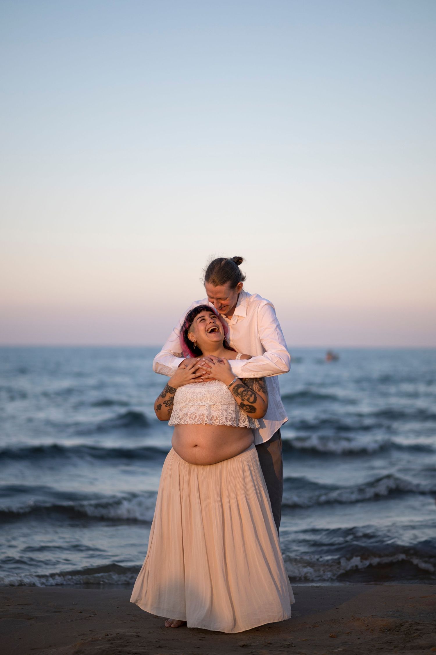 A man embraces his pregnant wife from behind as she laughs and smiles up at him. They stand facing the camera in front of Lake Michigan.
