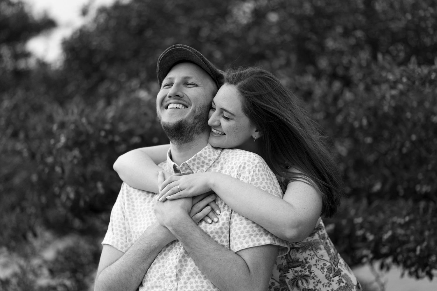 A b&w image of a woman wrapping her arms around her fiancé from behind while he clasps her hands on his chest and smiles.