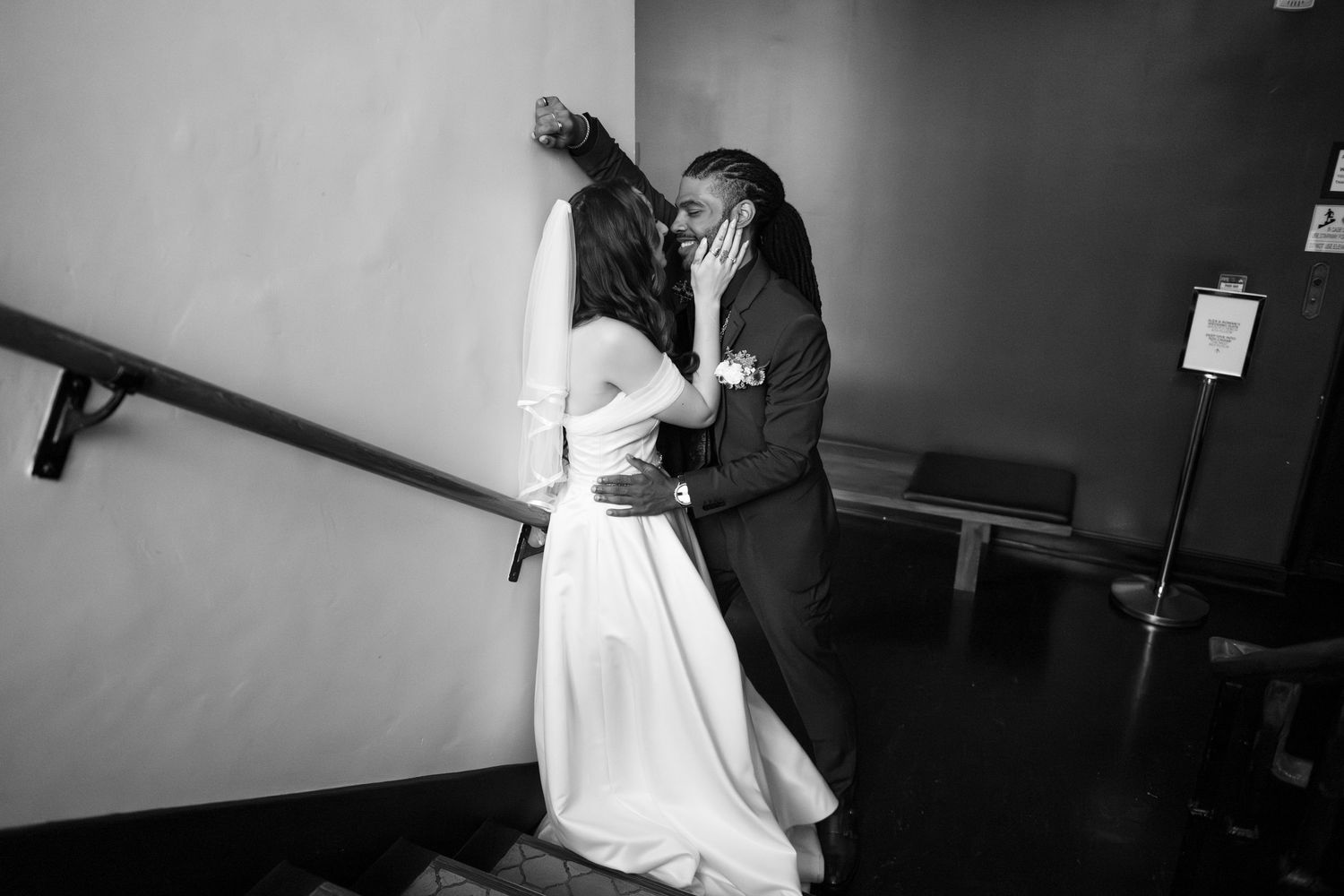 A groom kisses his bride against a wall - his hand on her waist, and her hand on his cheek (B&W).