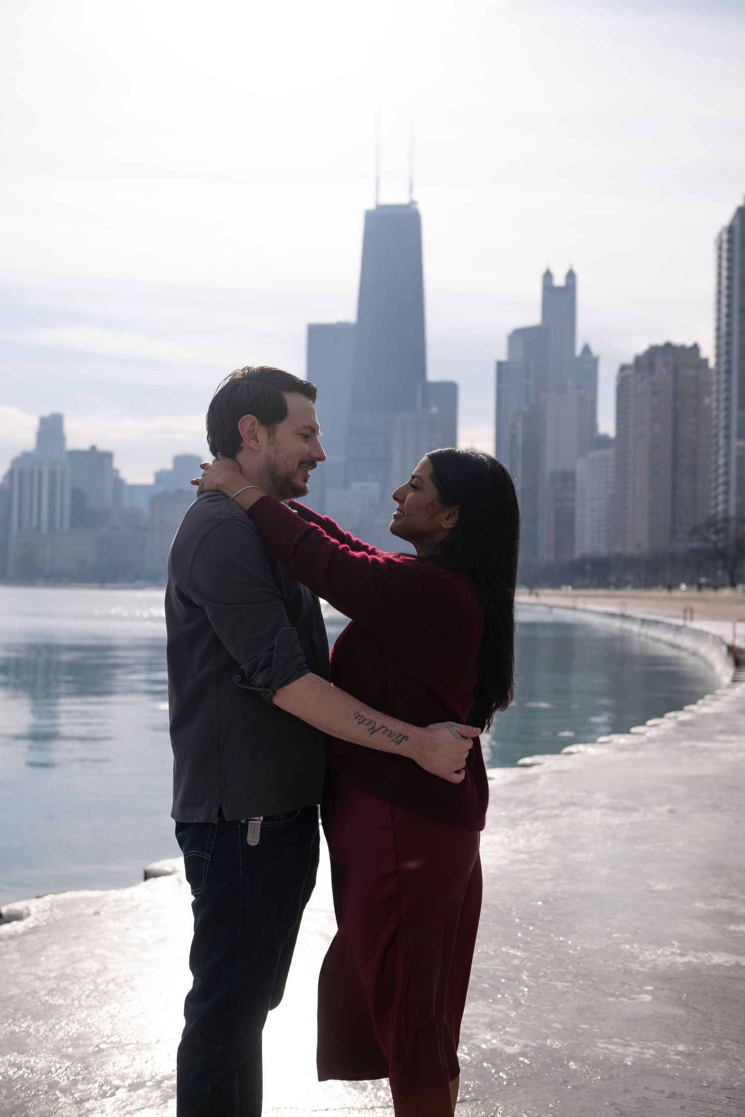 A cis-het couple stand facing and gazing at each other, arms wrapped around one another, with the Chicago skyline and lake in the background