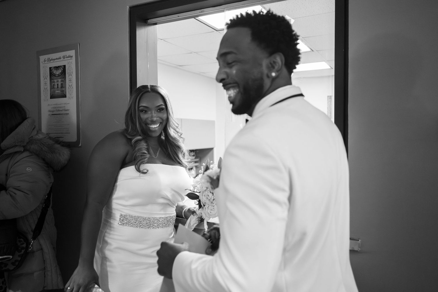 A couple stand at a reception window, smiling and laughing, while checking in for their city hall wedding (B&W).