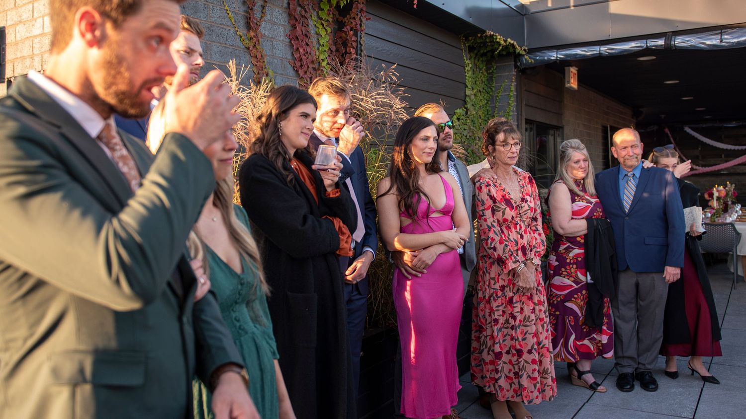 Family and friends stand in the shade and watch their loved ones get married at a rooftop wedding on a sunny day, while wiping away tears.