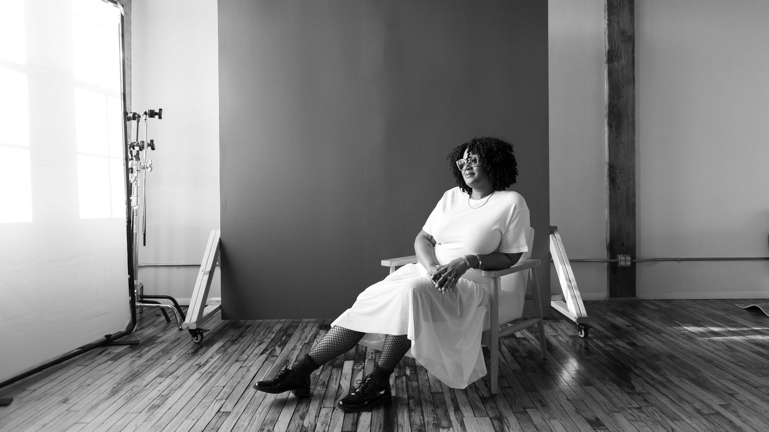 A woman with natural hair sits on a chair inside a photo studio in front of a backdrop, wearing all white and looking into the distance (BW)