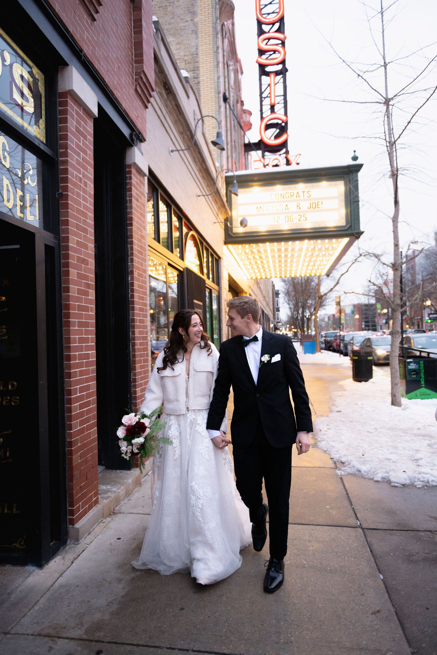 A young wedding couple hold hands and walk down the sidewalk past a theater marquee, smiling at each other, with snow on the ground.