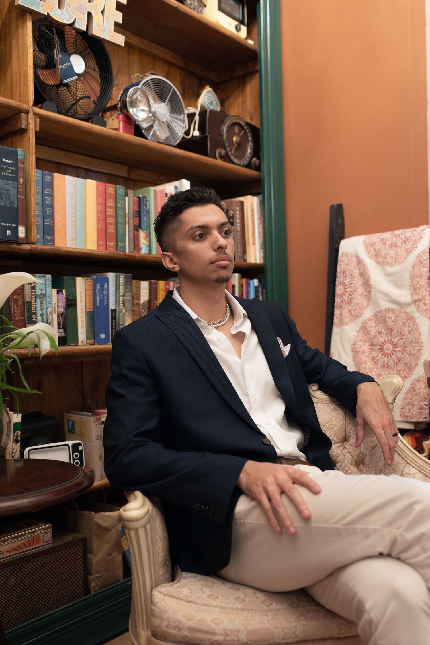 A newlywed groom sits on an armchair in front of a bookshelf, looking off into the distance.