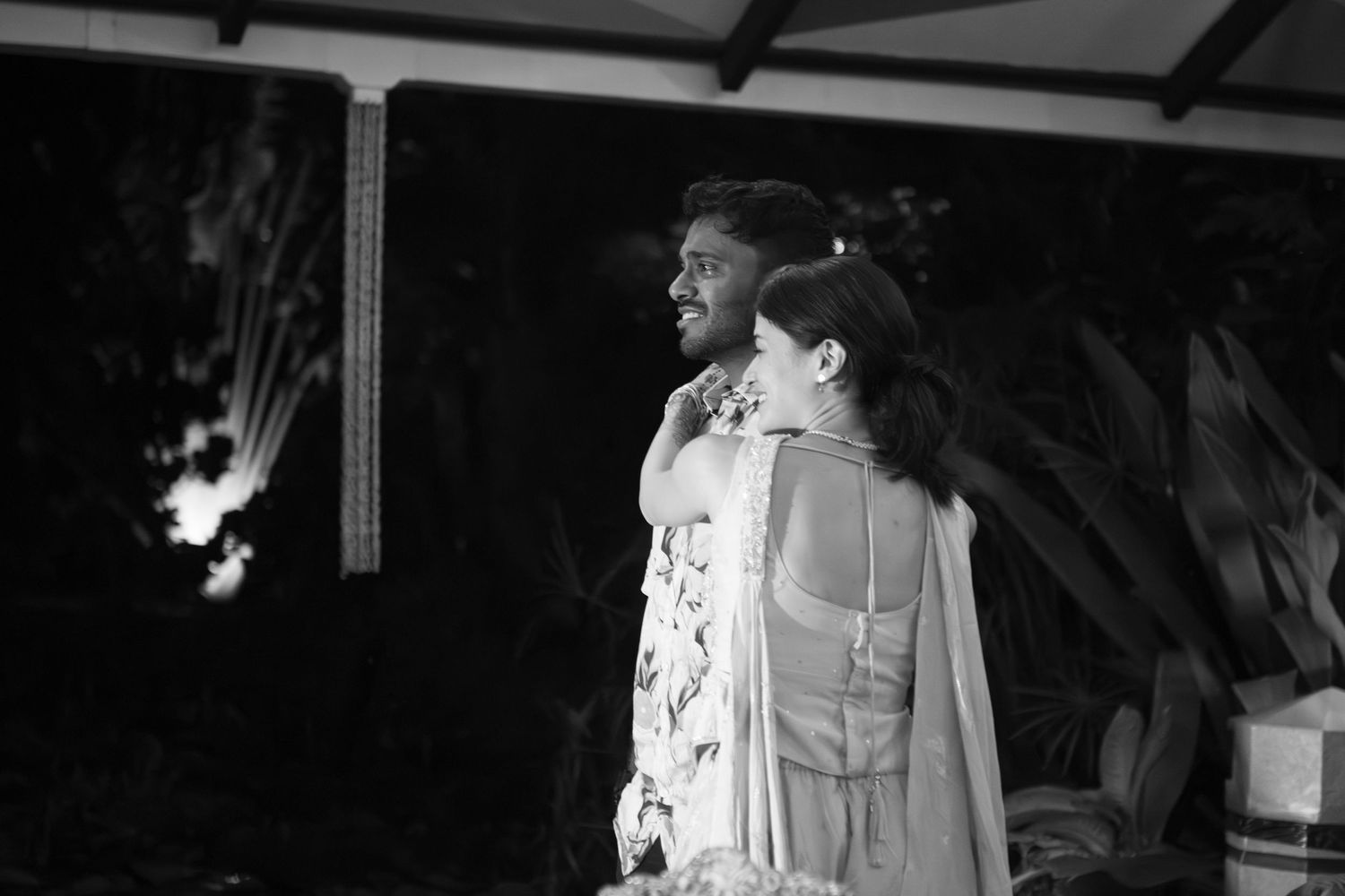 A wife hugs her husband as he cries and they both watch their beloved guests depart (B&W).