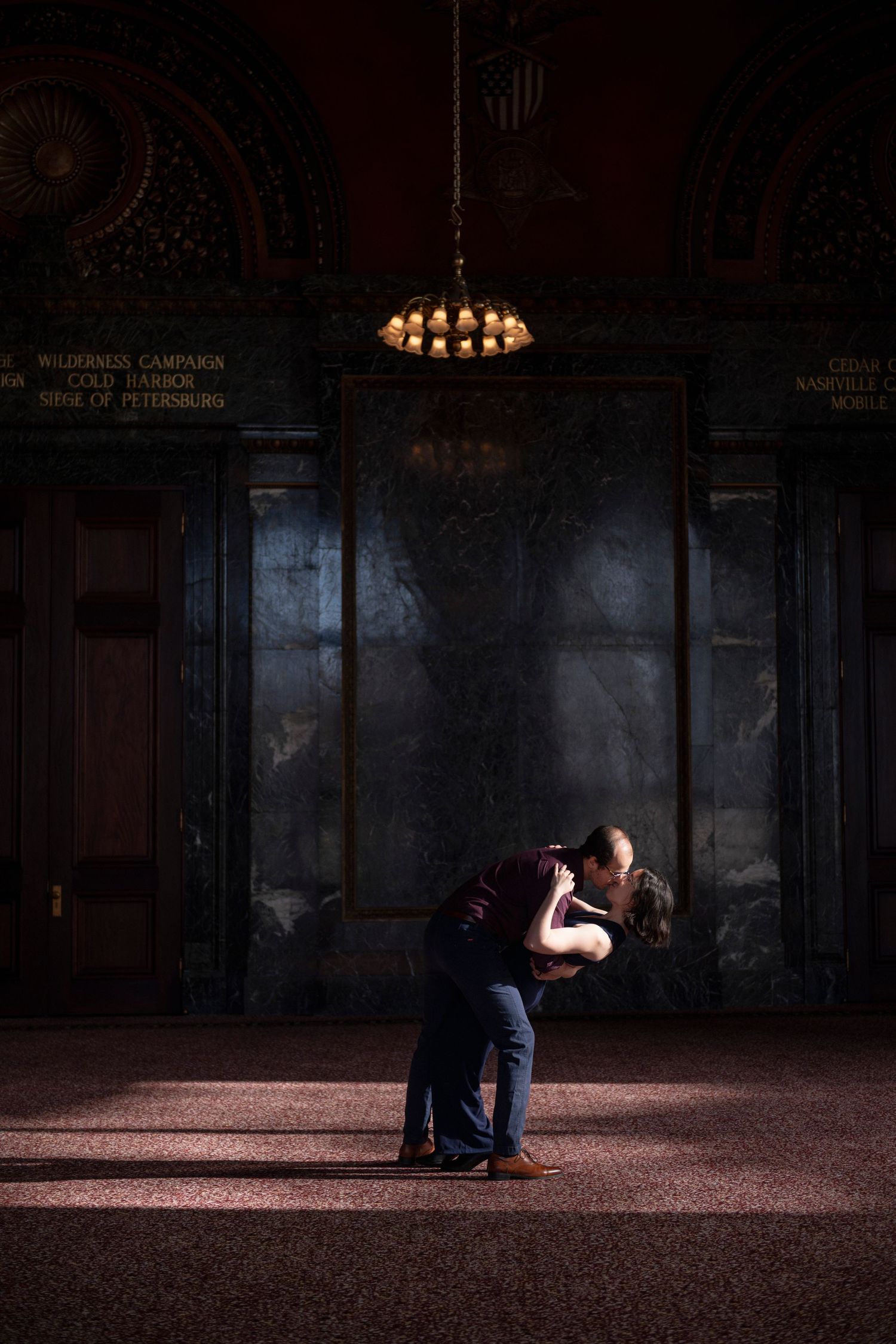 A newly engaged couple dip and kiss in a pocket of sunlight inside a moody hall of the Chicago Cultural Center.