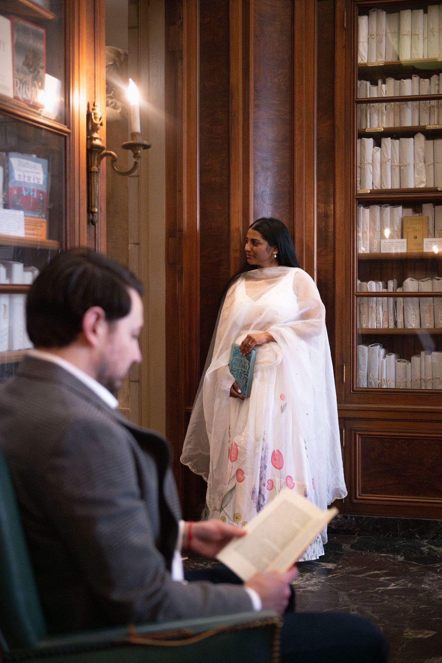 A man sits in the foreground, reading a book, while his fianceé leans against the doorframe of the library in the background.