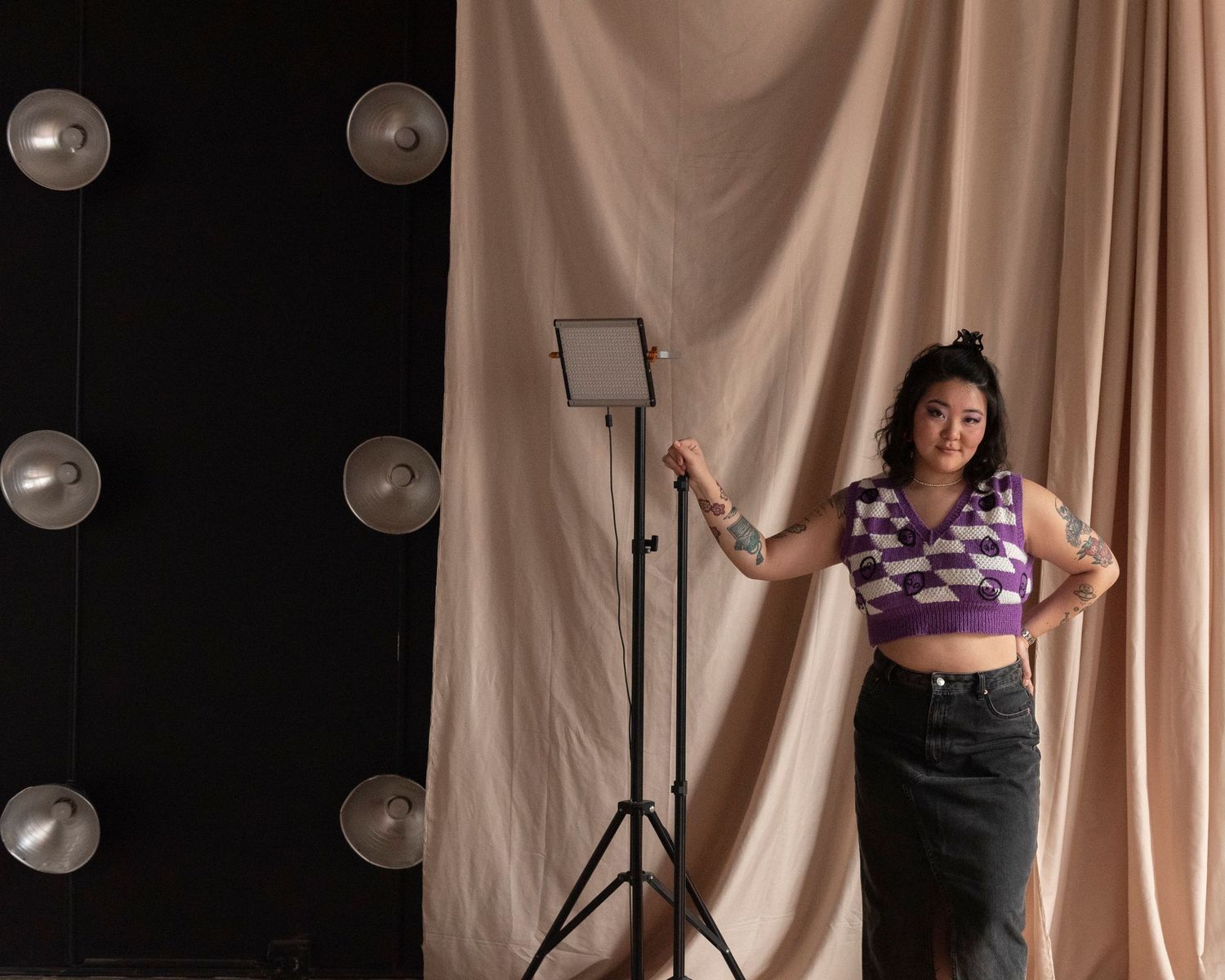 A young Asian woman photographer poses in a studio next to lighting equipment in a sweater vest and denim skirt.