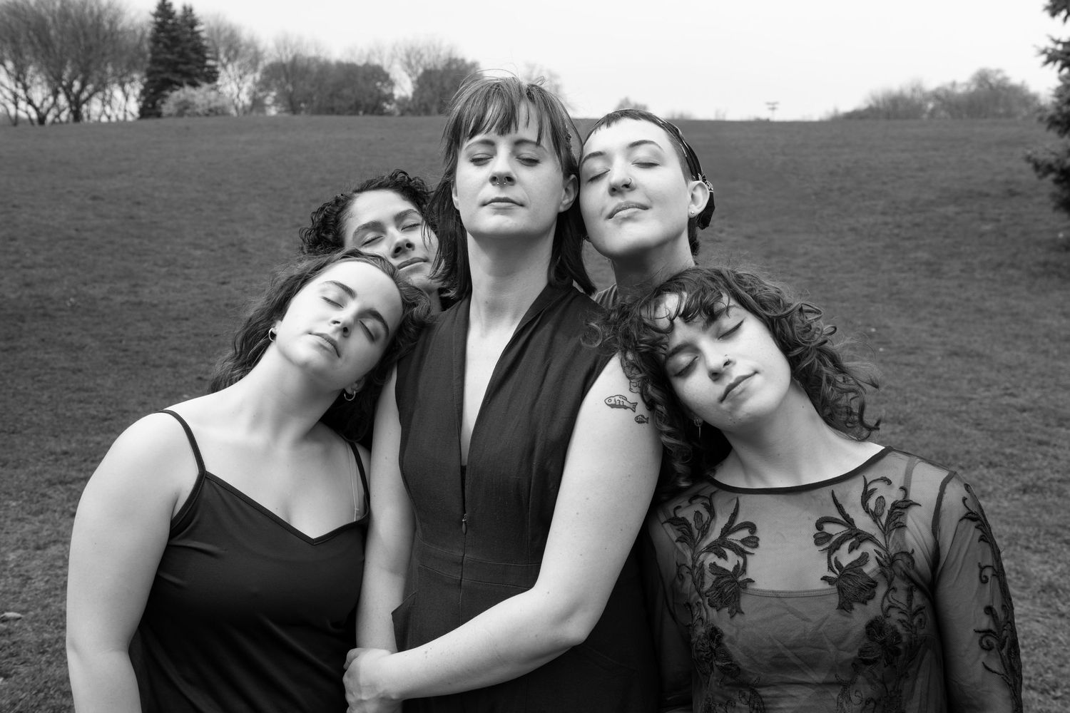A group of 5 femmes stand close, resting their heads together with faces upturned and eyes closed, in black and white.