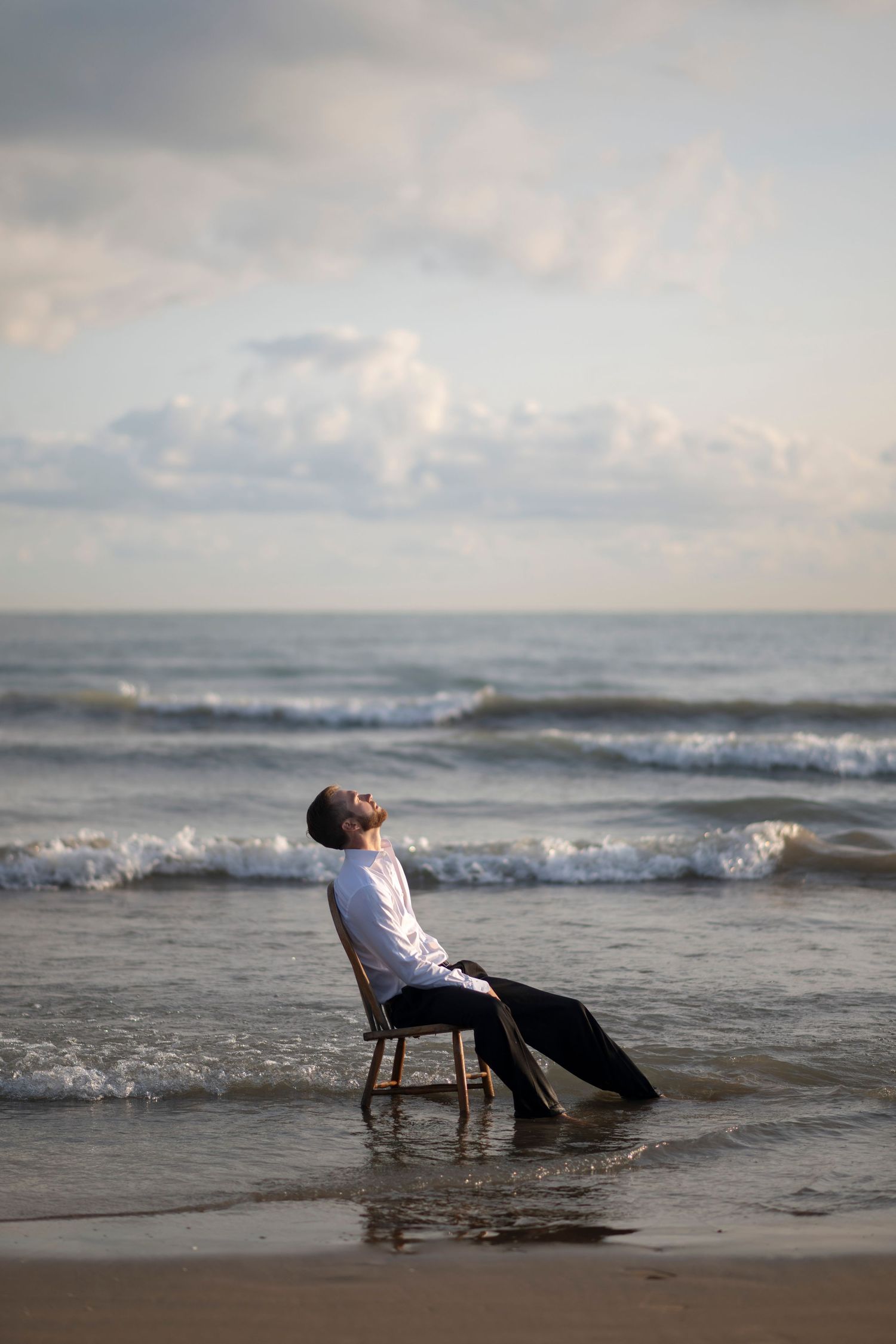 A businessman sits on a chair on the beach among shallow waves on the shores of Lake Michigan, resting and looking up into the sky.
