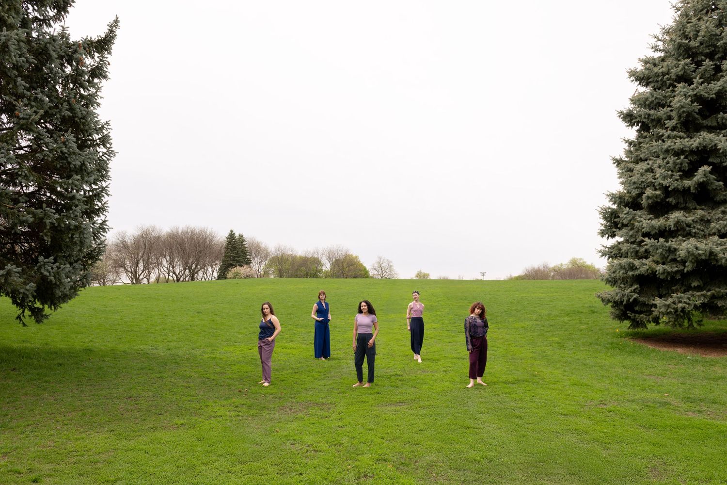 A group of 5 dancers stand on a hill, evenly spaced from each other, with a tree framing the group on either side, at Chicago's Horner Park.