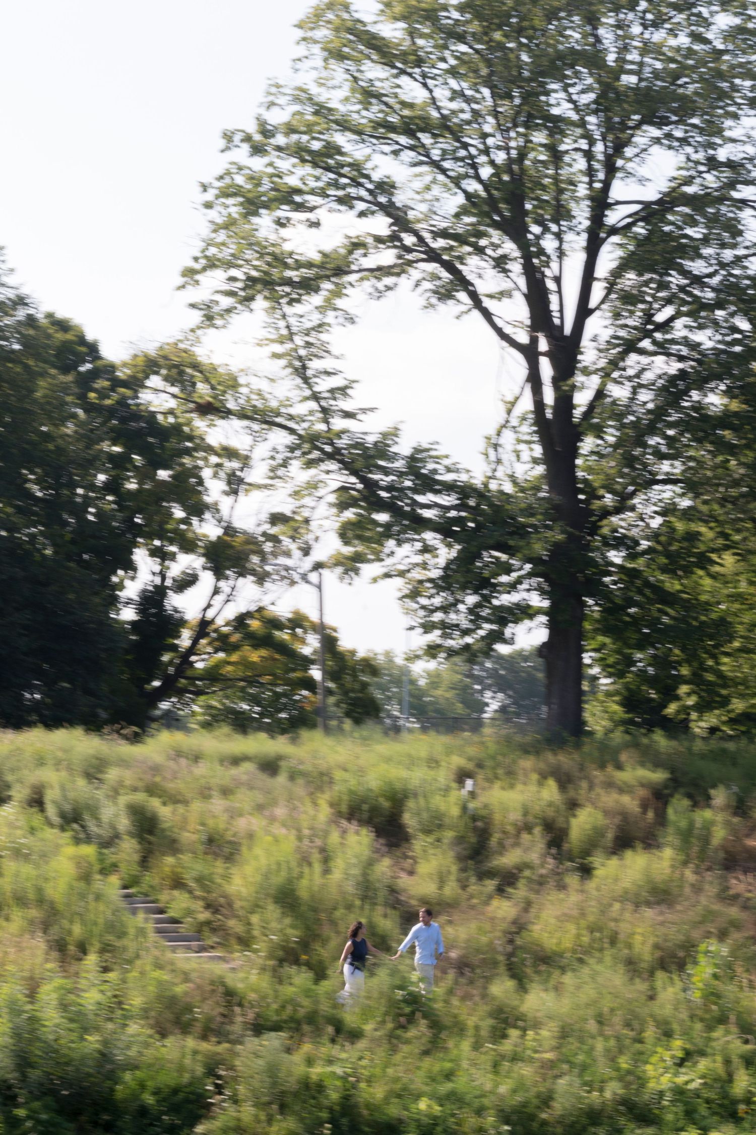 A couple hold hands and run across a grassy hill with trees in the background and motion blur.