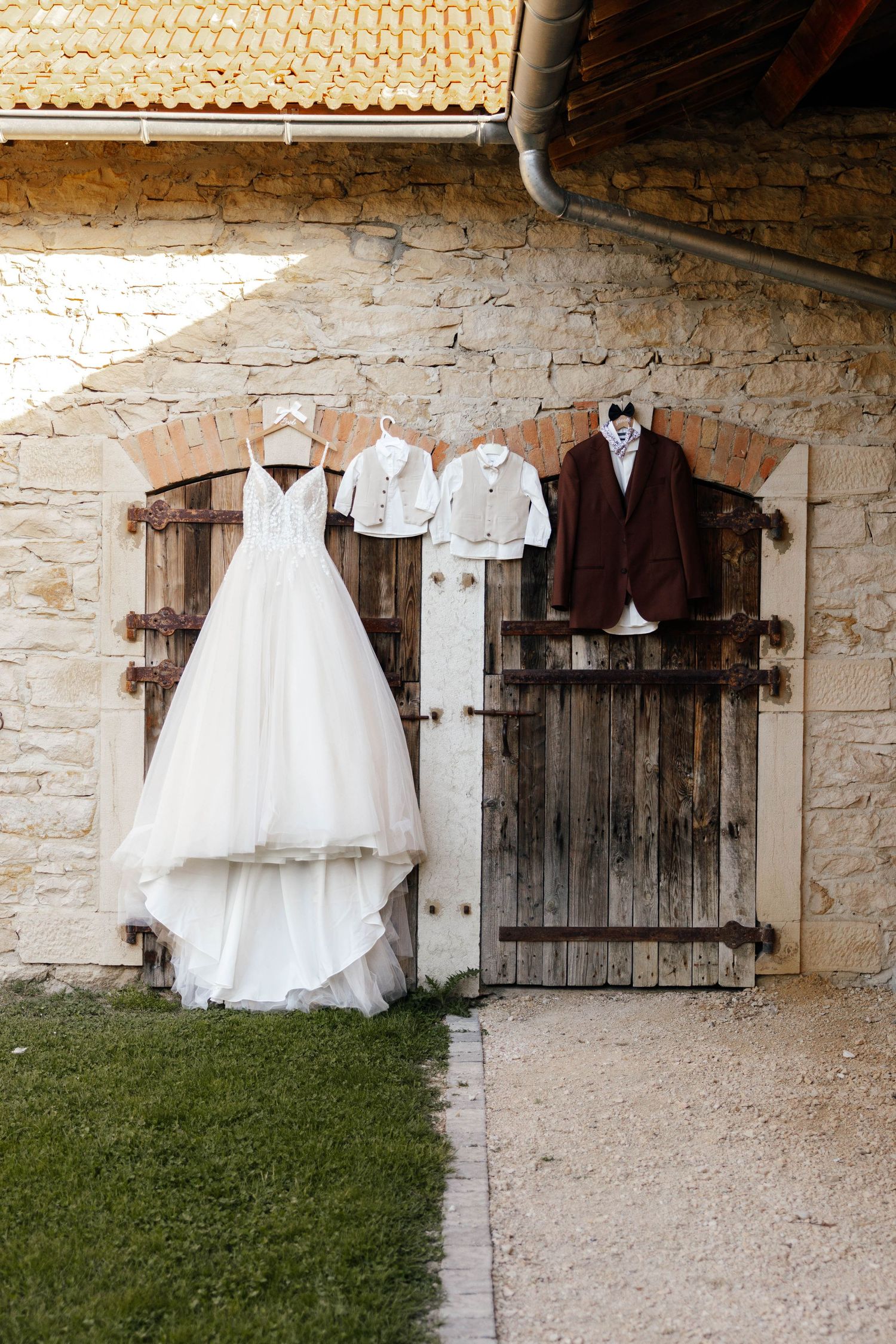 White bridal gown and childrens wedding outfits hanging on rustic wooden shutters of a stone building.